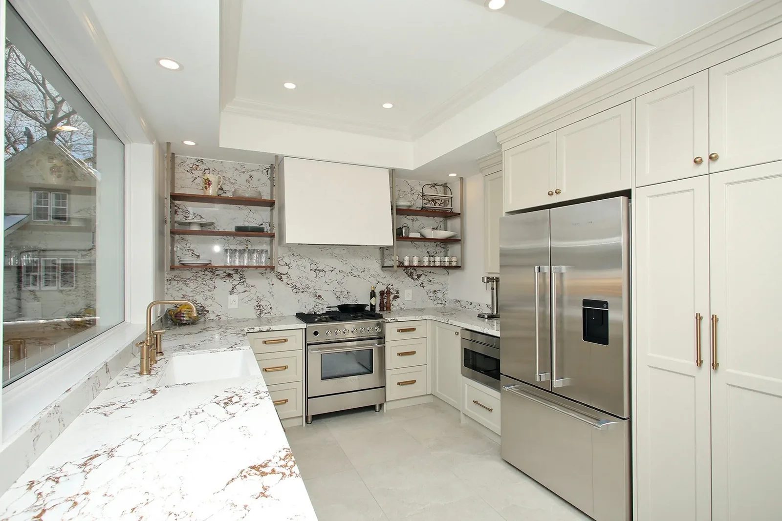 Bright white kitchen with veined stone counters, brass hardware, open shelves and stainless appliances.