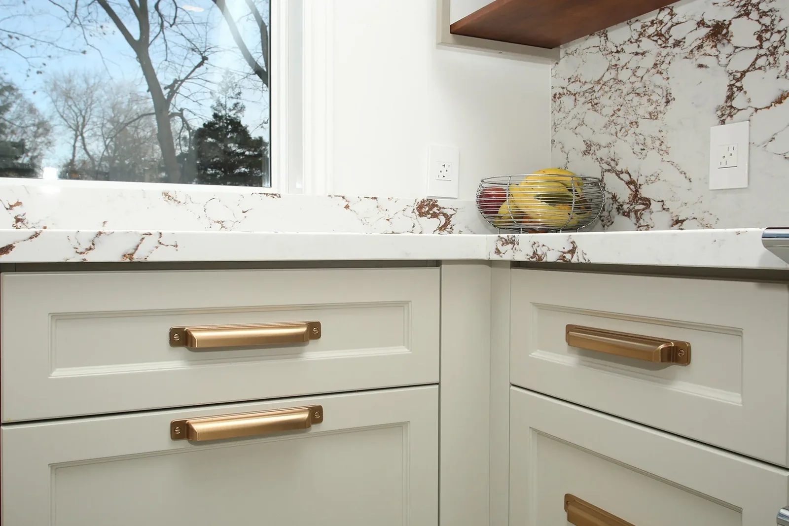 Warm white kitchen drawers with brass pulls and bold veined quartz beside a bright window.