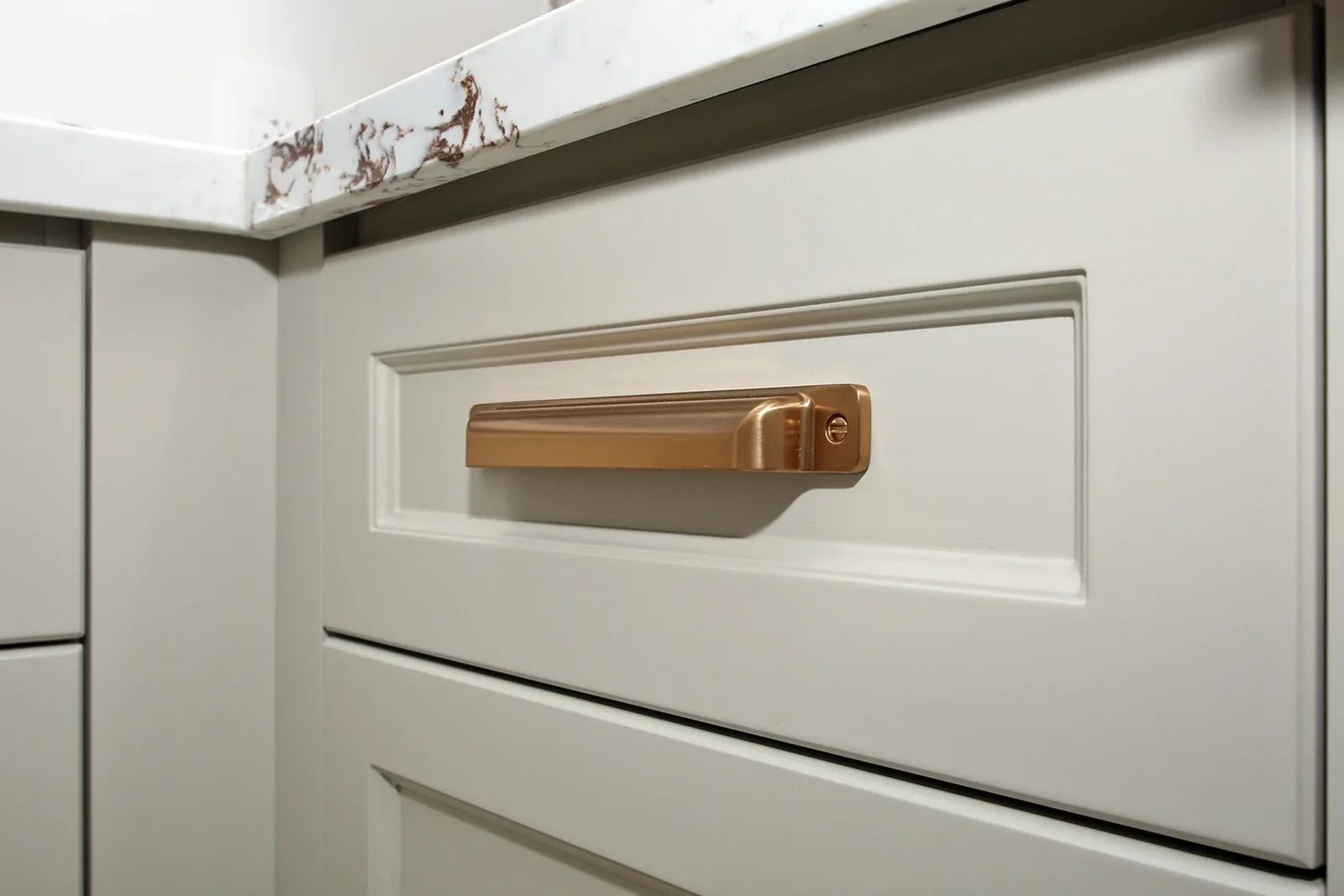 Warm white shaker kitchen drawers with a copper-toned pull and quartz countertop in a finished renovation.