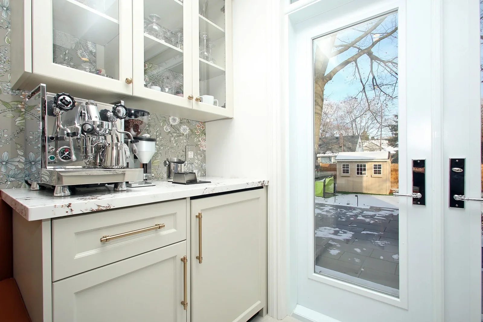 Bright white coffee bar with brass pulls, quartz counter, floral backsplash, and glazed door to the yard.