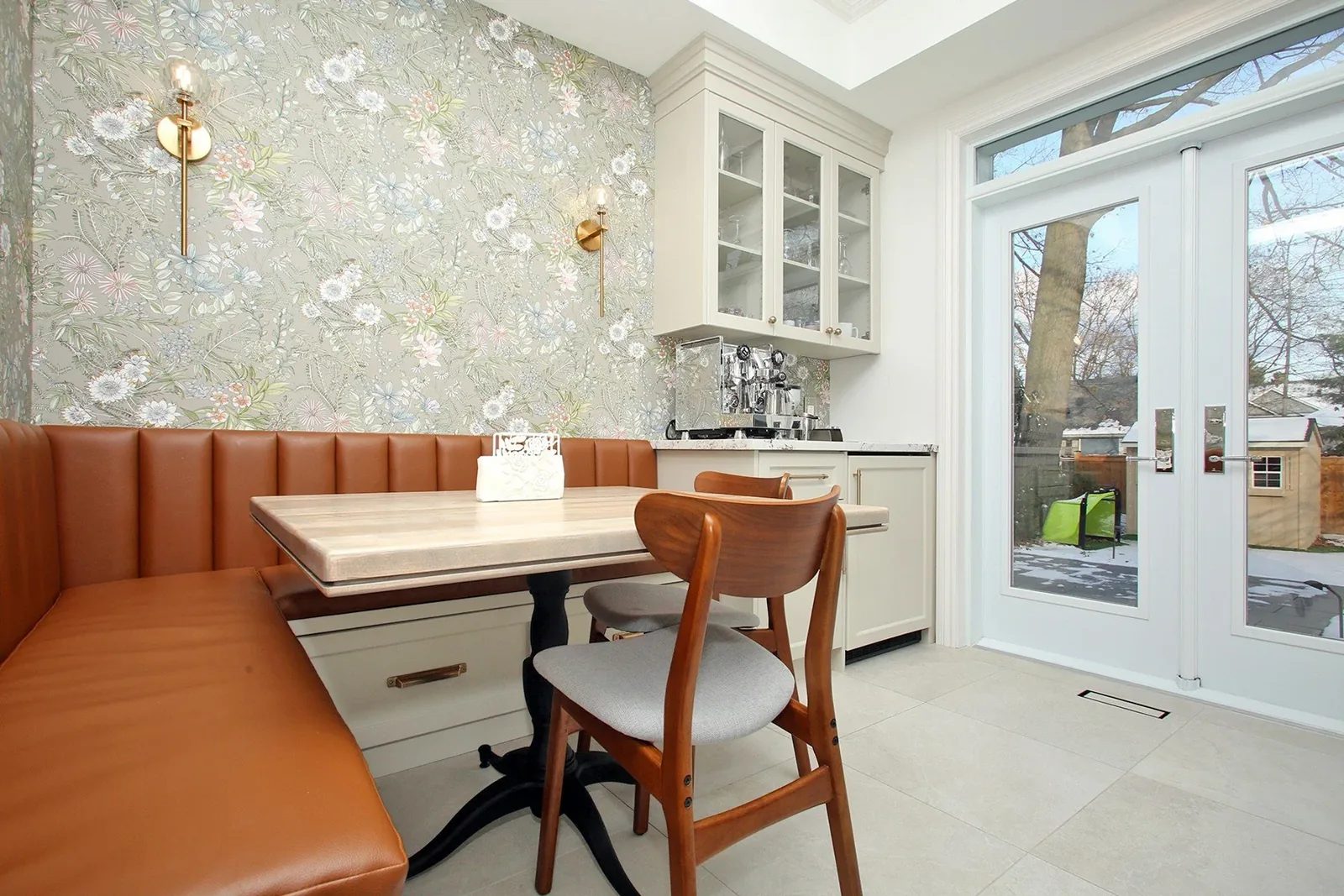 Bright kitchen breakfast nook with a caramel banquette, floral wallpaper, brass sconces, and garden doors.