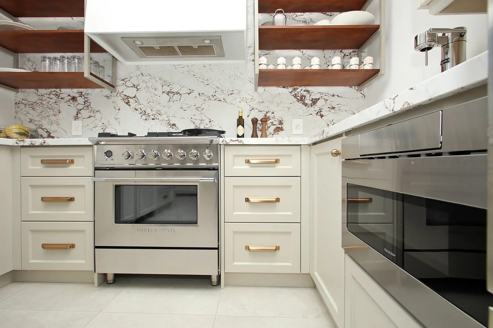 Warm white kitchen with brass pulls, veined stone backsplash, open wood shelving and stainless appliances.