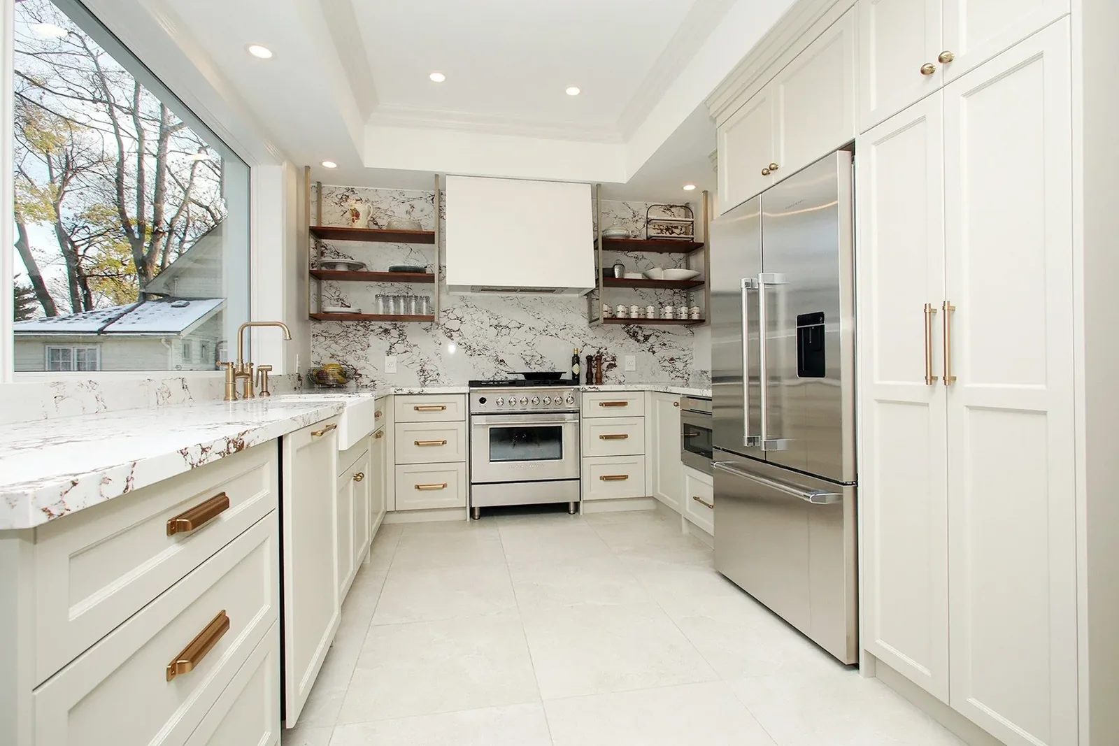 Bright white kitchen with brass hardware, marble look surfaces, and a large window over the sink.