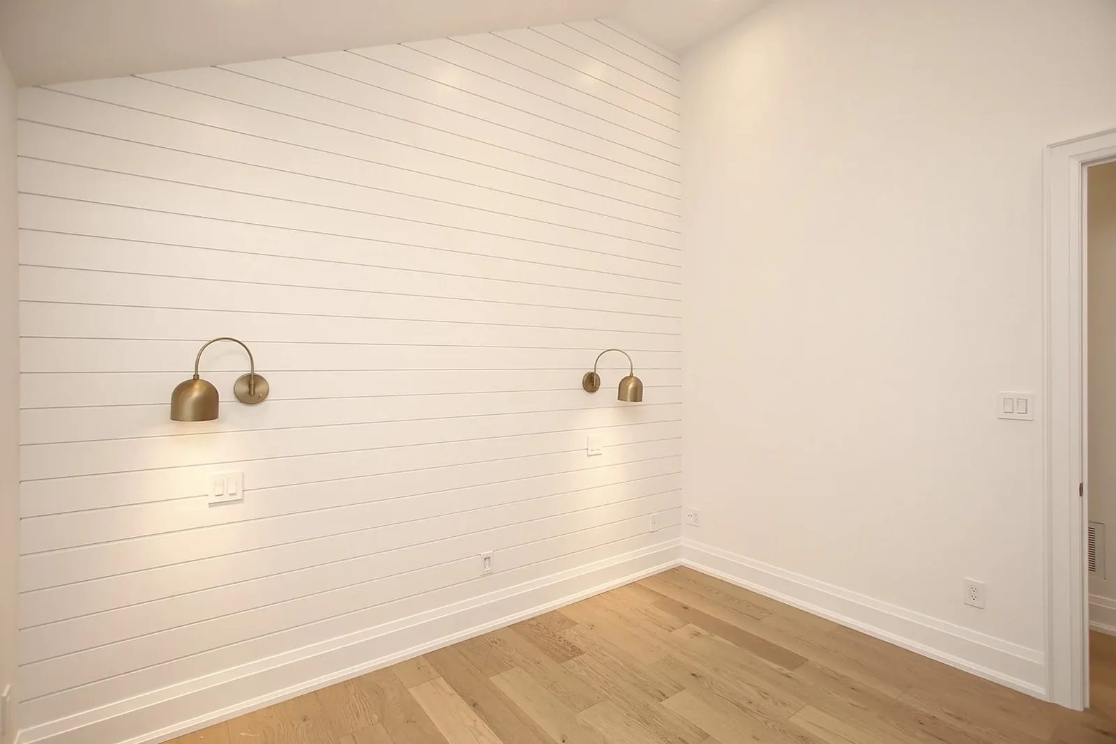 Bedroom corner highlighting a white shiplap wall with two brushed brass sconces above oak hardwood flooring.