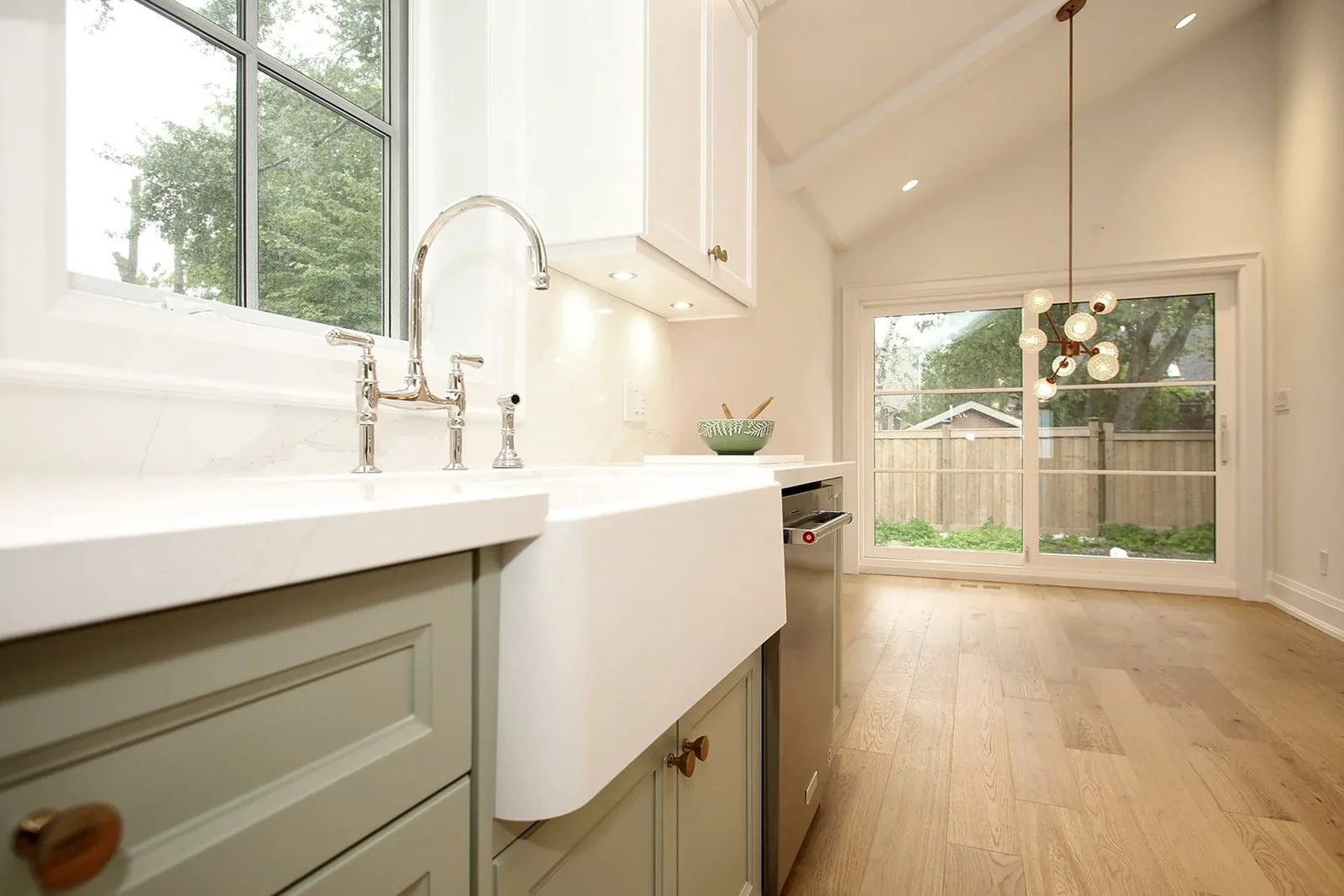 Apron sink and chrome bridge faucet at a large window with a vaulted ceiling and a bubble chandelier beyond in the nook.