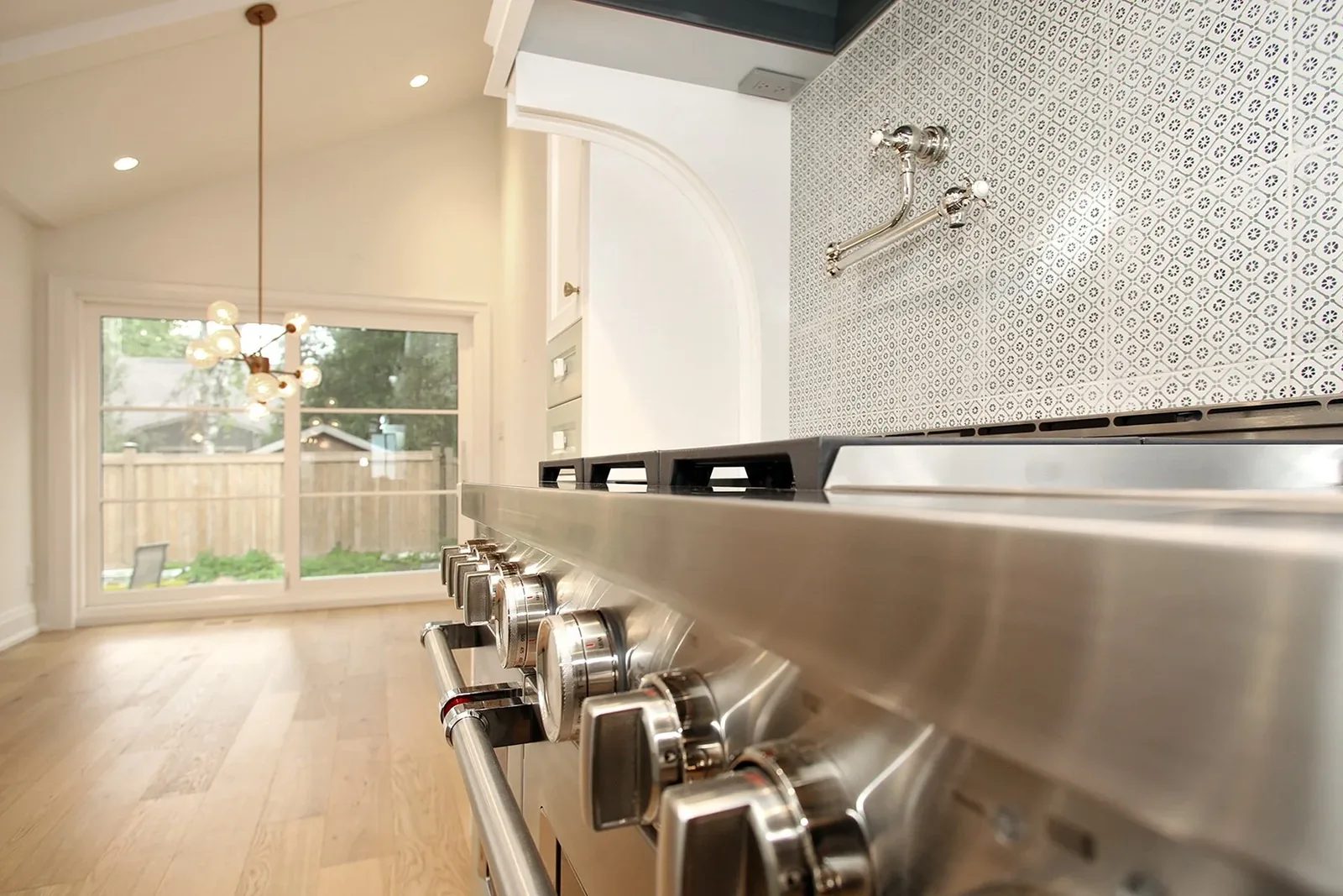 Close perspective along a stainless pro range toward a dining nook lit by a brass globe chandelier in a vaulted space.