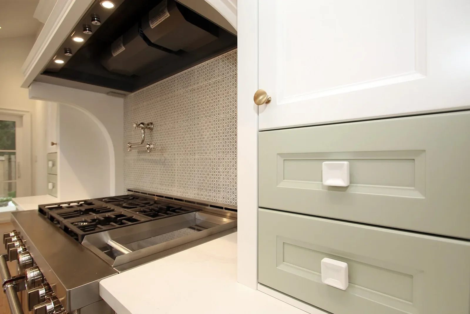 Detail of sage green inset drawers beside a stainless pro range and patterned geometric backsplash under a custom hood.