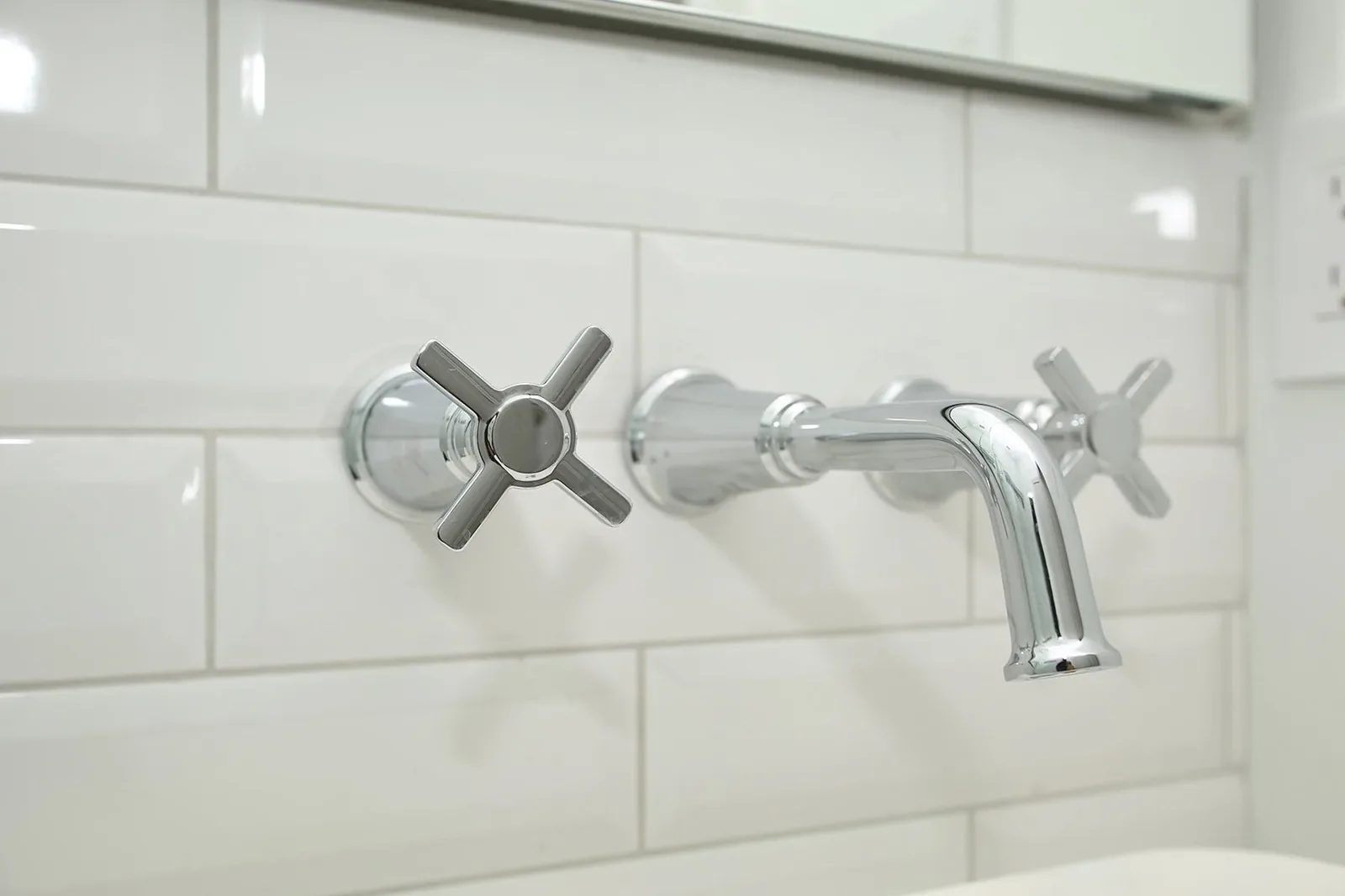 Close up of a polished chrome wall mount bathroom faucet with cross handles on white beveled subway tile.