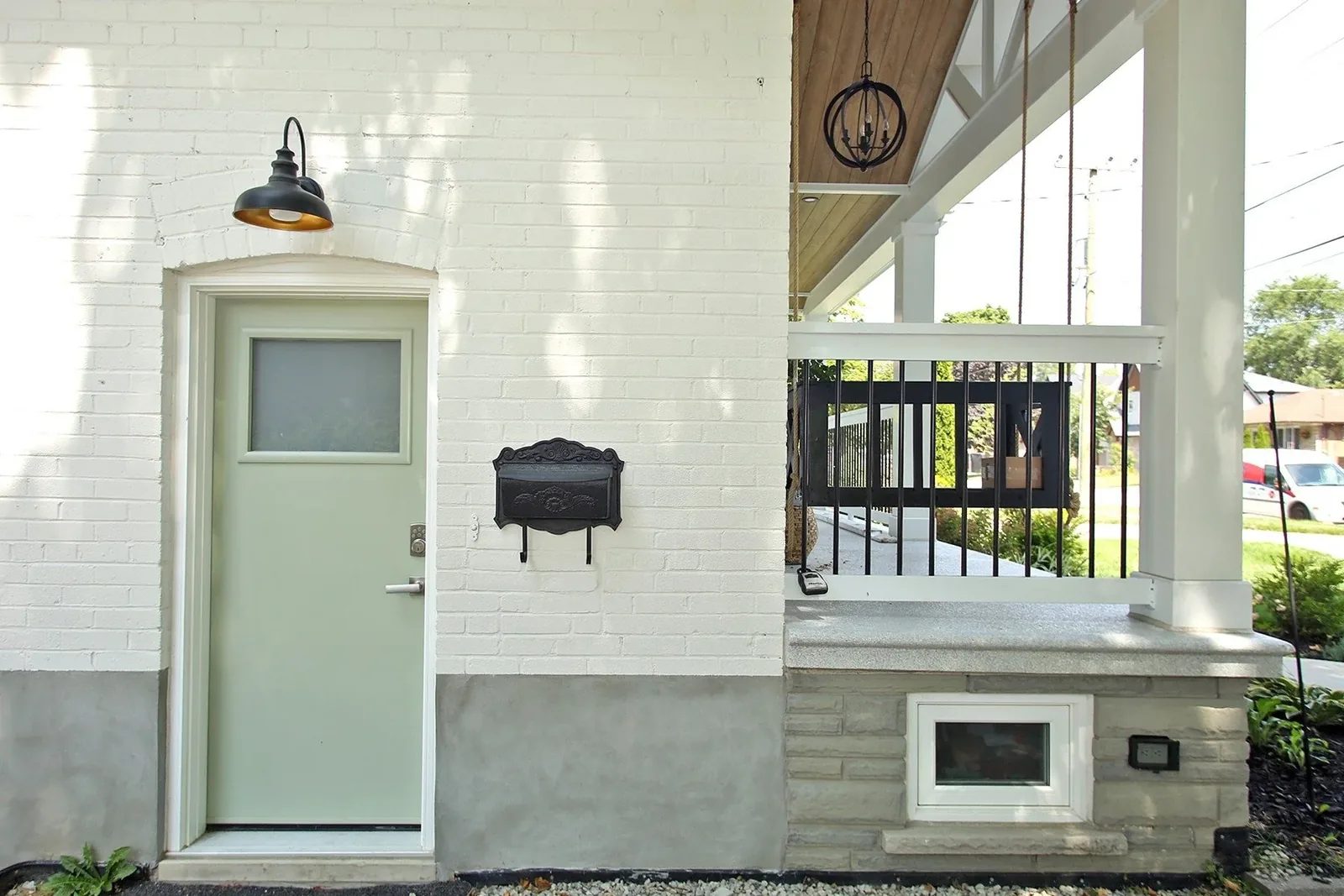 Painted white brick side entry with a sage green door, black barn sconce, cast iron mailbox, and a stone skirt below.