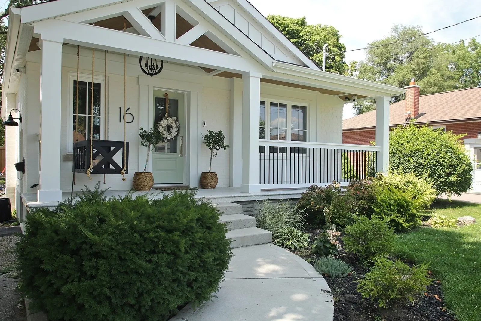 Front porch of the white bungalow with a sage green door, wreath, house number sixteen, and a timber truss gable above.