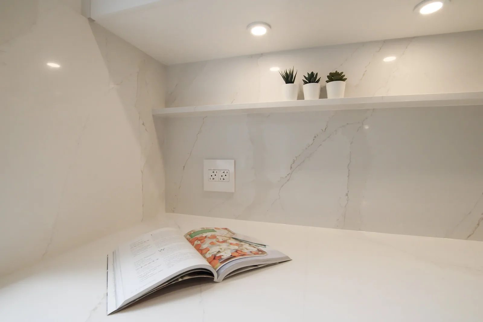 Warm white quartz counter niche with marble look wall panels, recessed lighting and a slim display shelf.
