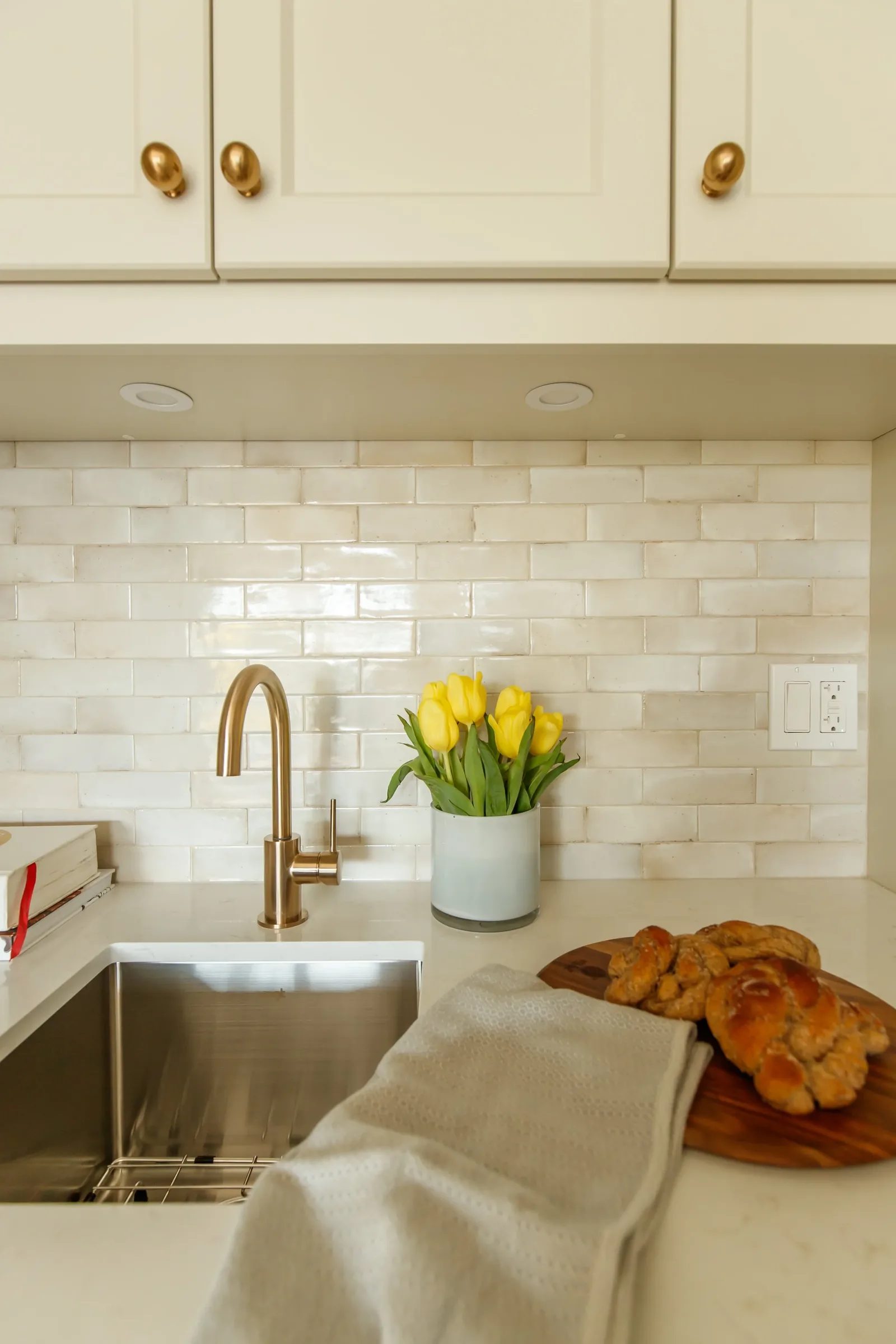Kitchen vignette with brass gooseneck faucet, stainless apron sink, tulips and challah on a wood board.