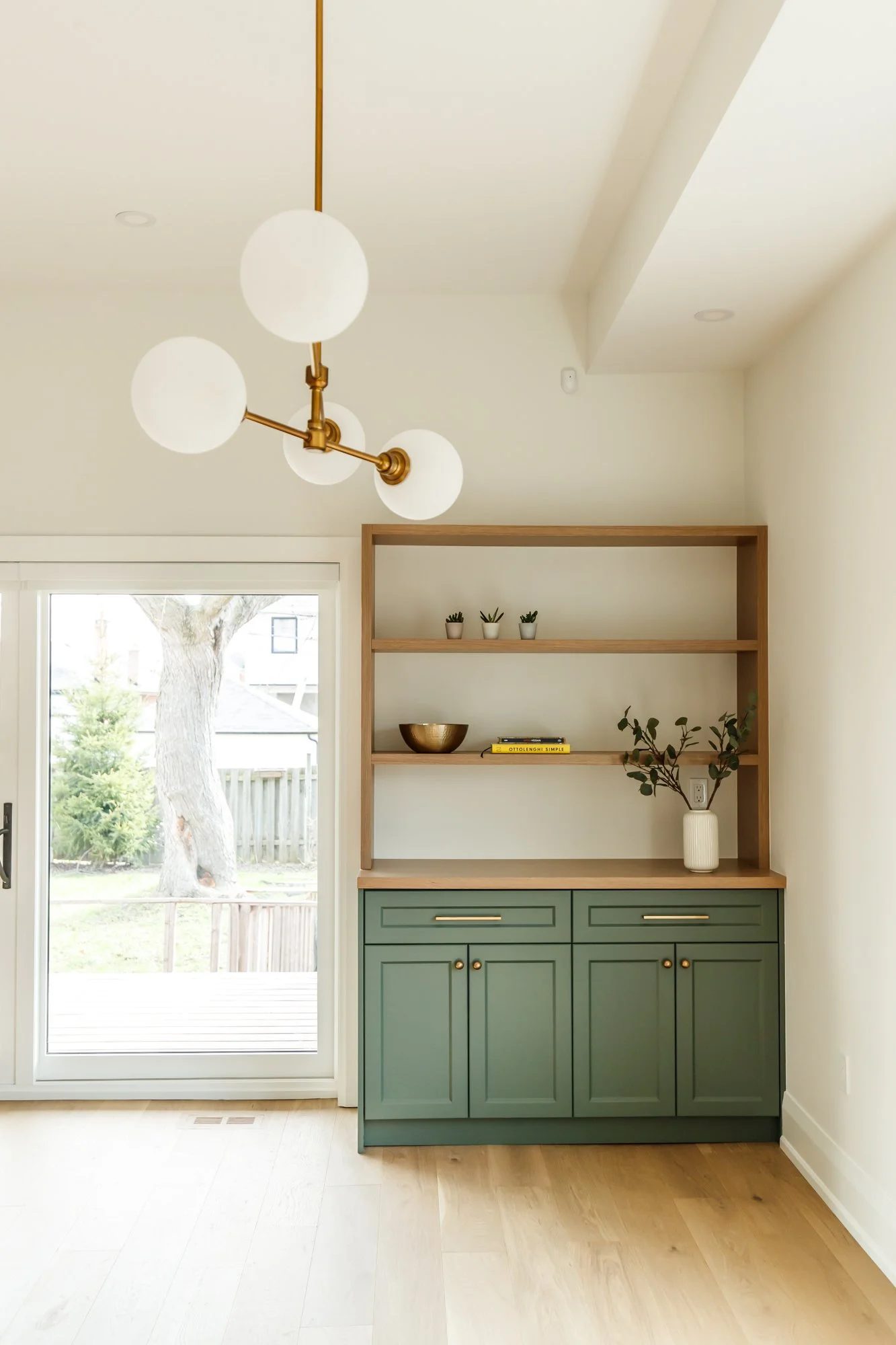 Sage green built-in cabinetry with oak shelving, brass hardware, and a globe chandelier in a bright dining area.