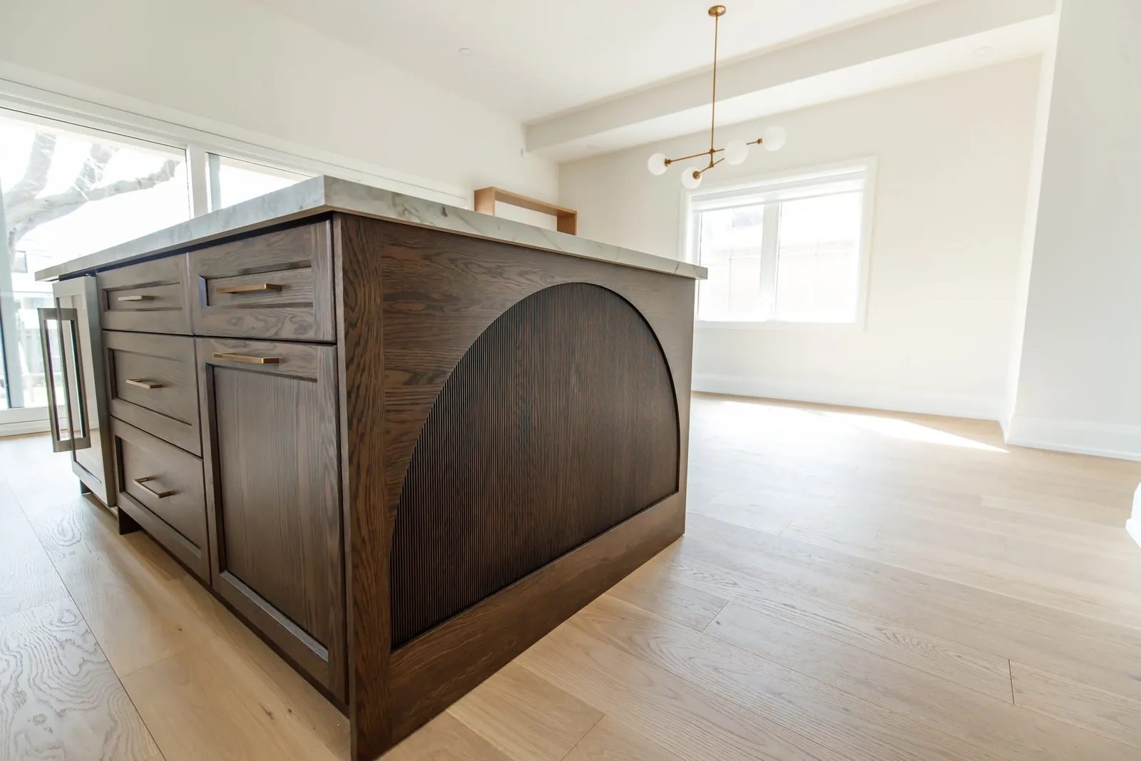 Close angle of a stained oak island with an arched fluted end panel beside a bright dining zone.
