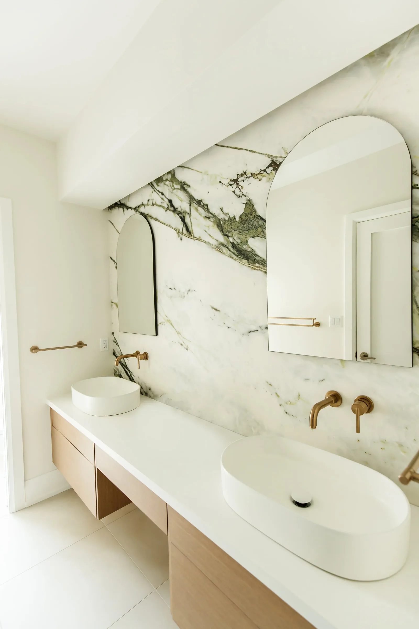 Modern bathroom vanity with marble slab wall, brass fixtures, oak drawers and twin white basins.
