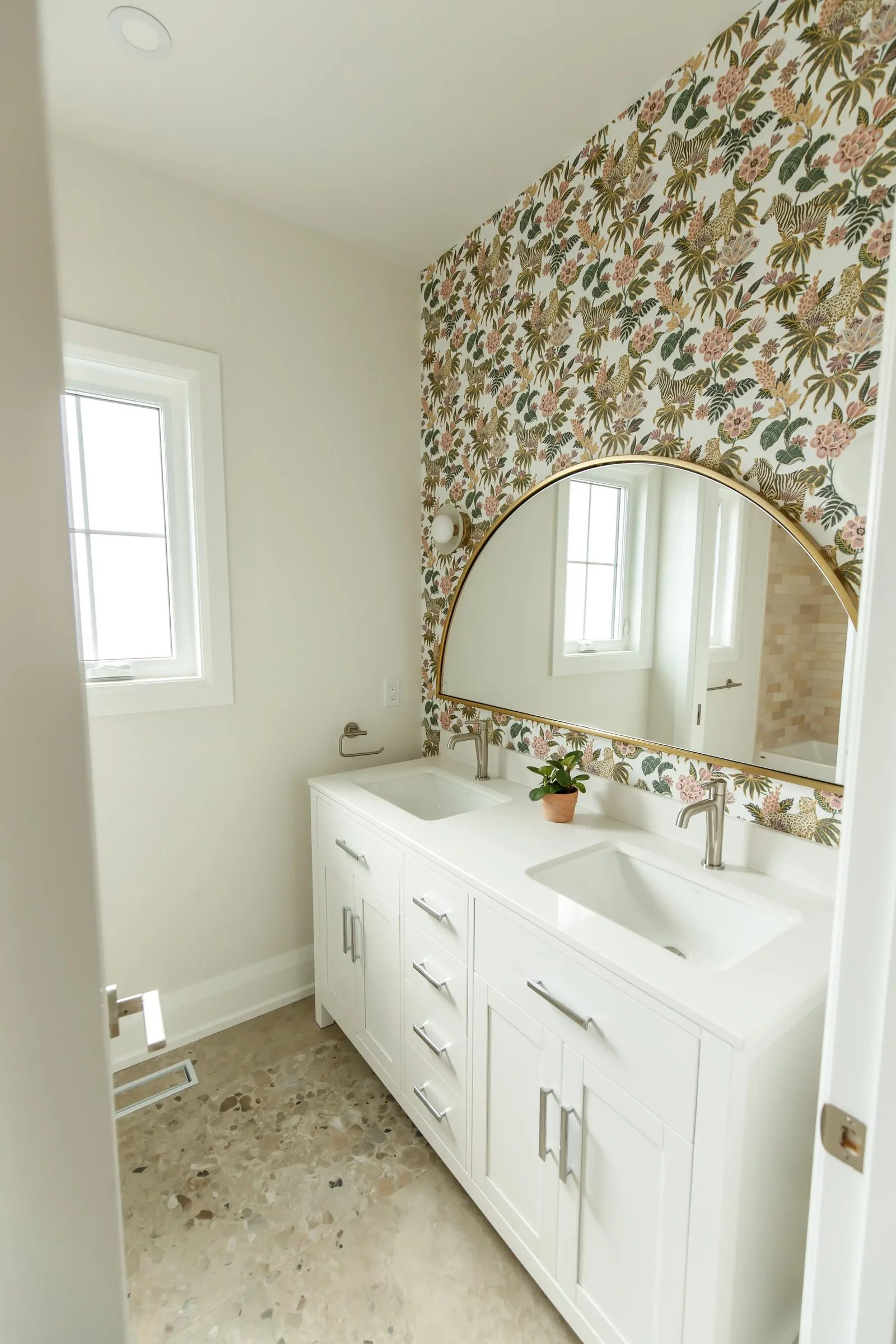Naturally lit double-vanity bathroom with arched gold mirror and floral wallpaper accent wall.