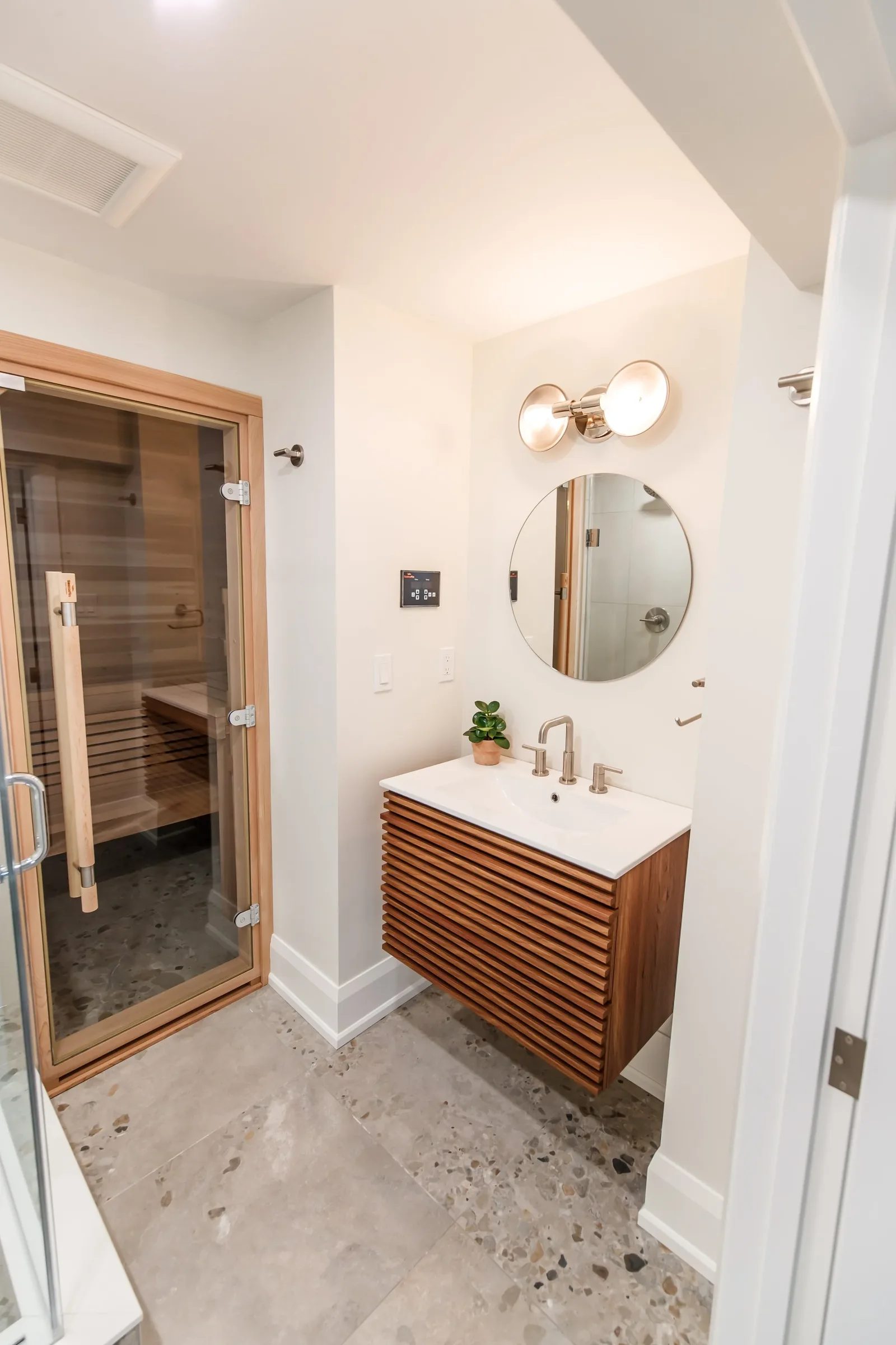 Spa-style basement bathroom with cedar sauna, slatted walnut floating vanity and round mirror above terrazzo tile floor.