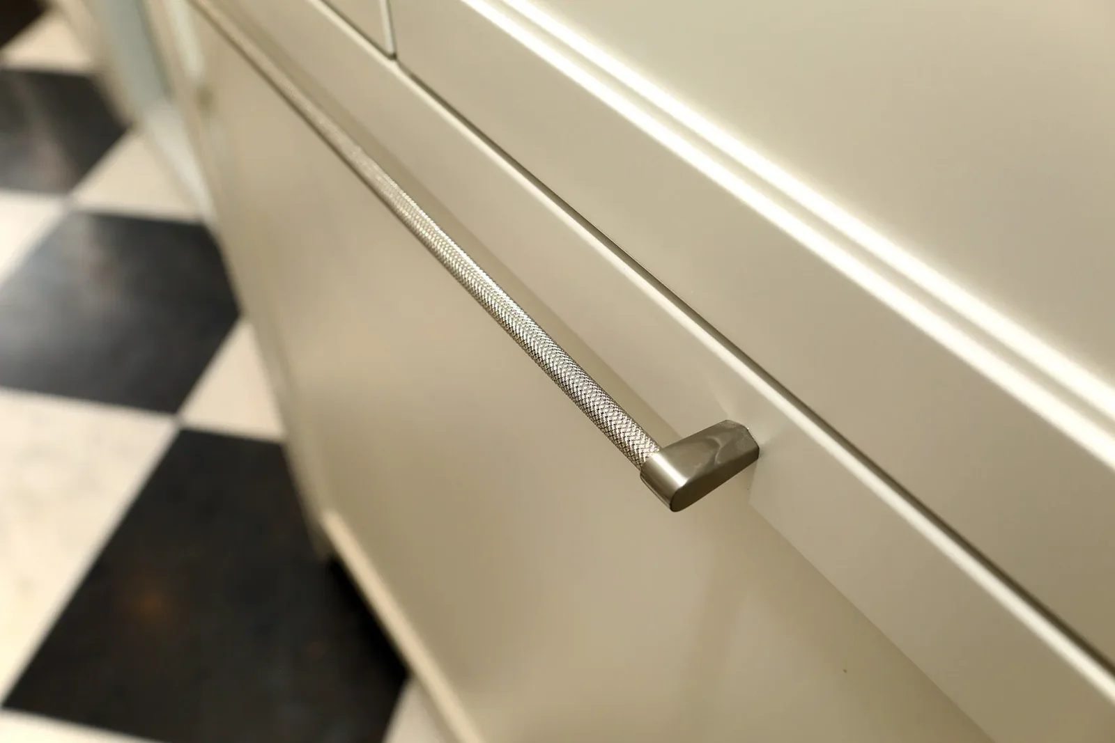 Cream kitchen cabinetry with a textured nickel pull and black and white tile flooring in soft light.