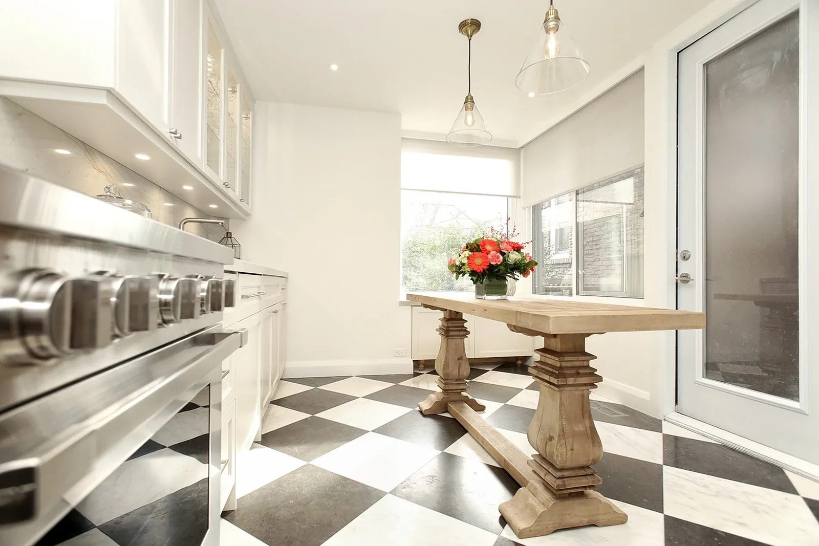 Bright white kitchen with checkerboard floor, trestle table, pendant lights and a large window.