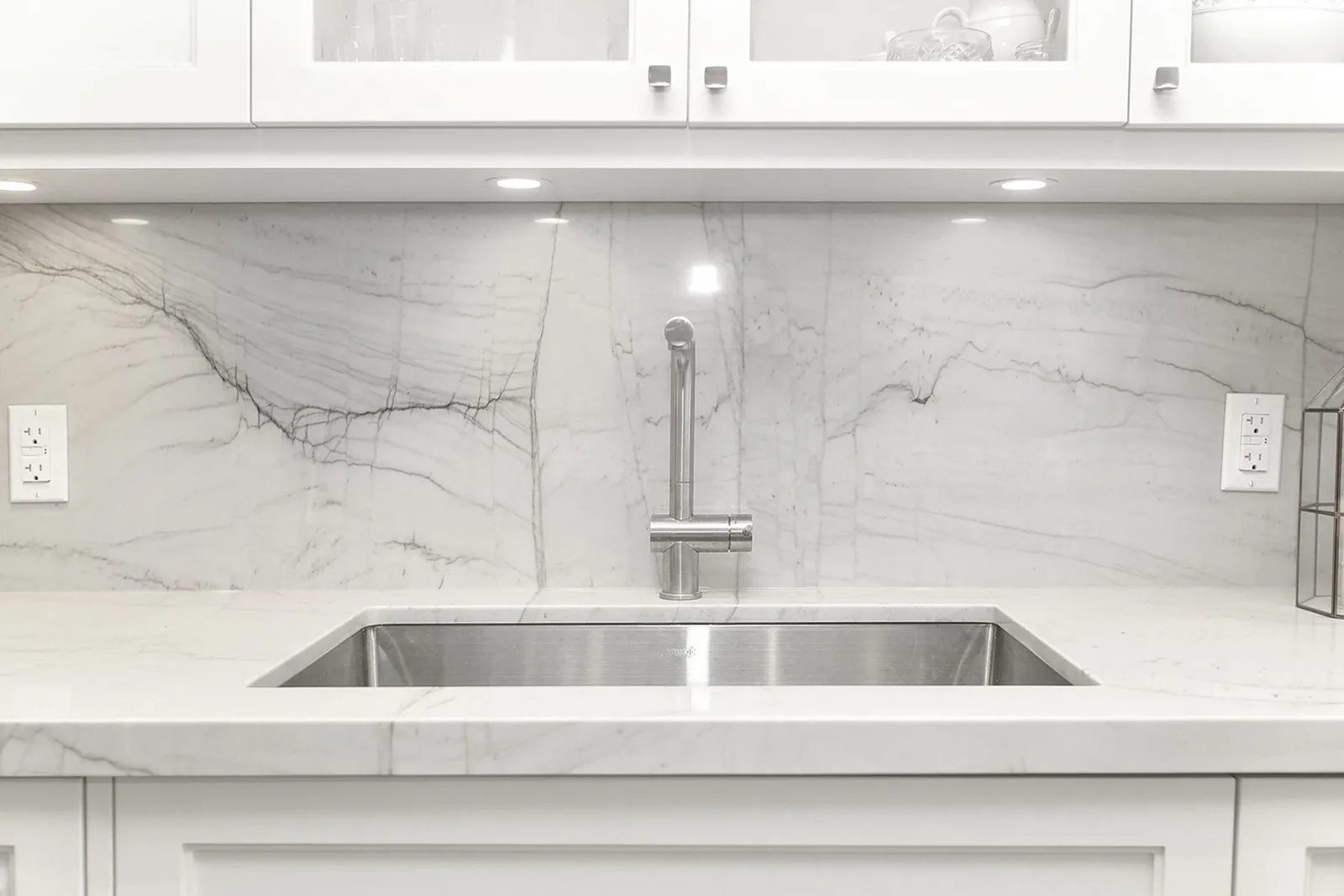 White kitchen sink wall with veined stone backsplash, stainless fixtures, and clean modern millwork.
