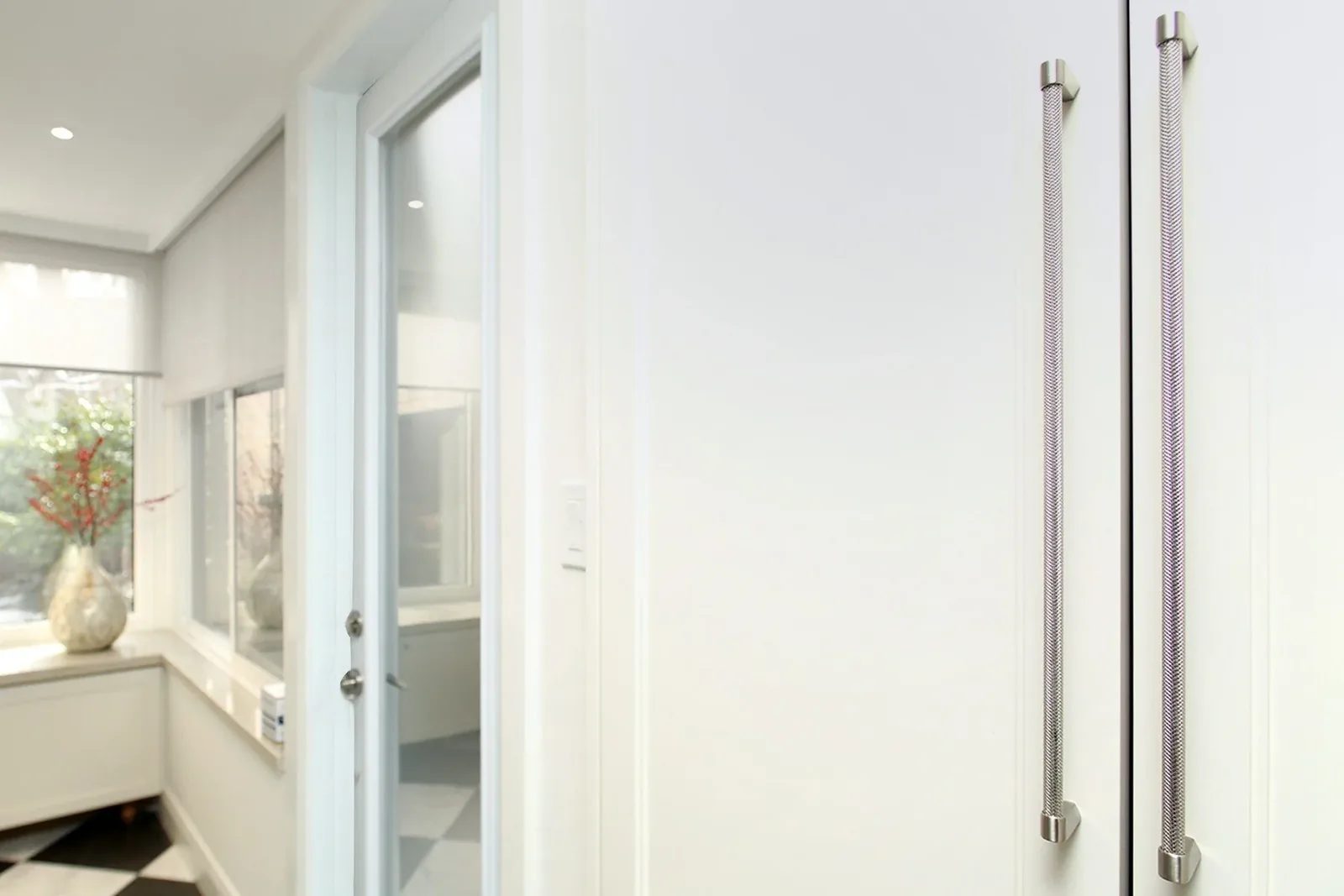 Bright white hallway with a glass door, tall brushed metal pulls, and checkerboard tile by the windows.