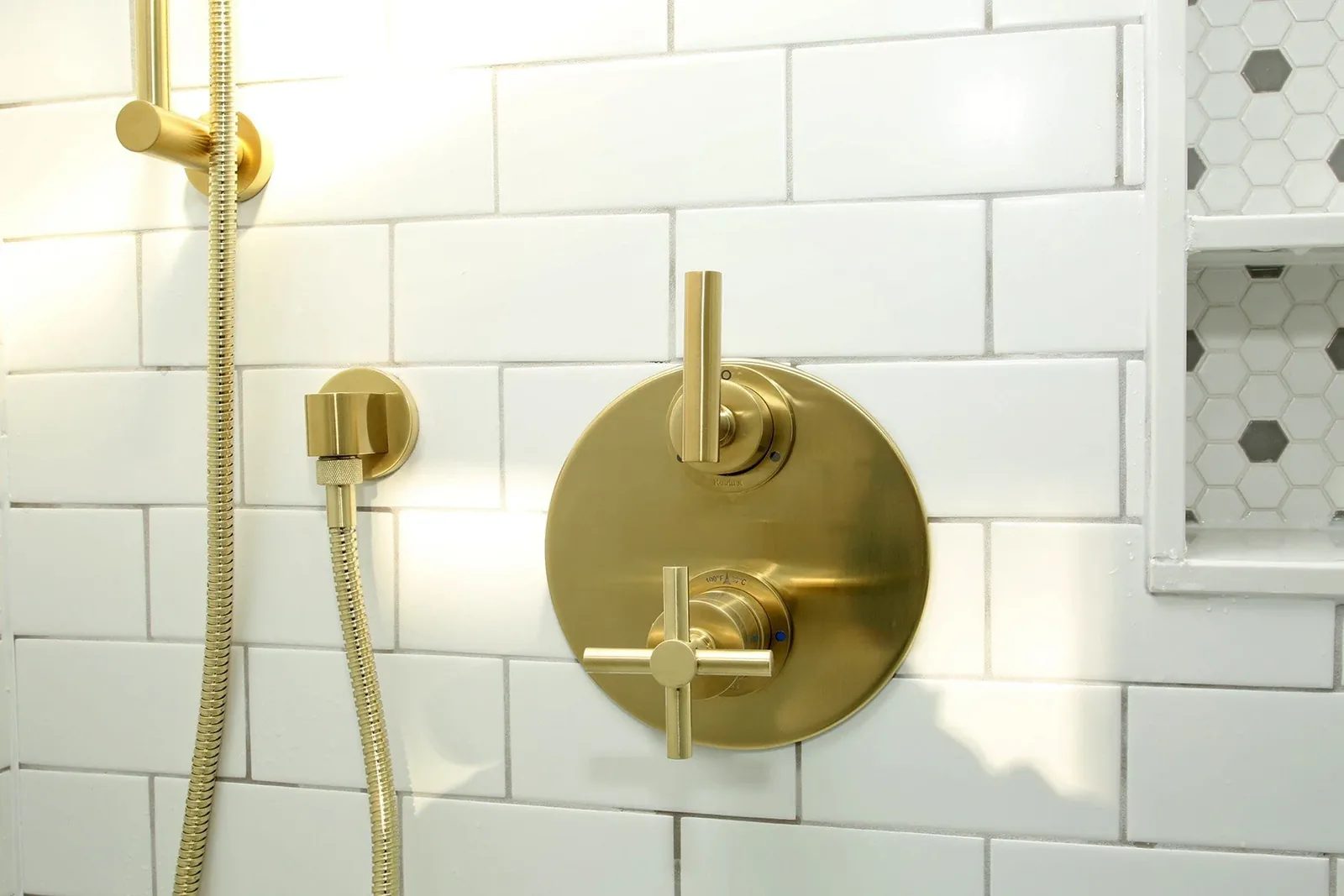 Brushed brass shower controls set against warm white subway tile with a tiled niche in the corner.