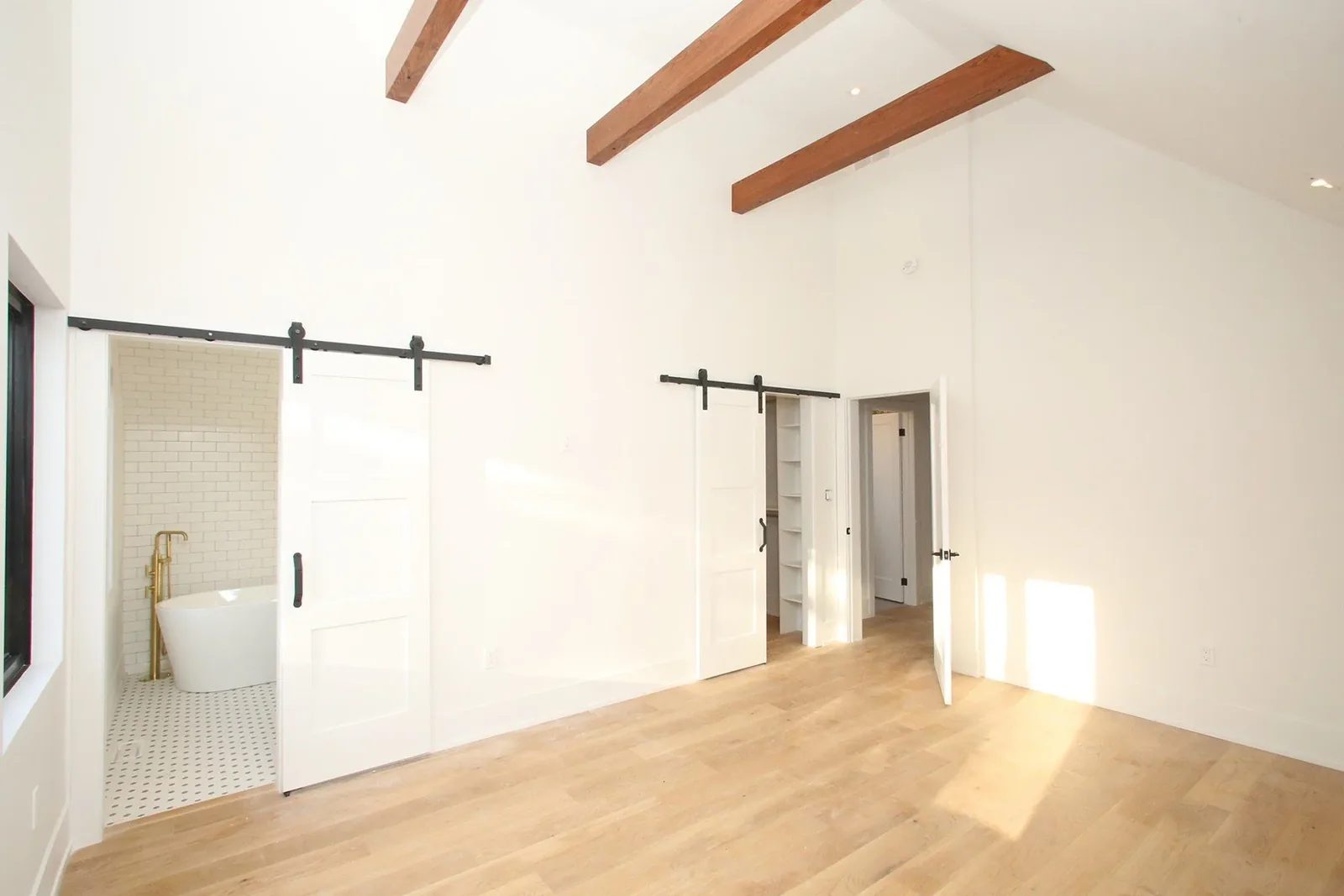 Bright finished primary bedroom with oak floors, exposed beams, barn doors, ensuite tub, and closet access.