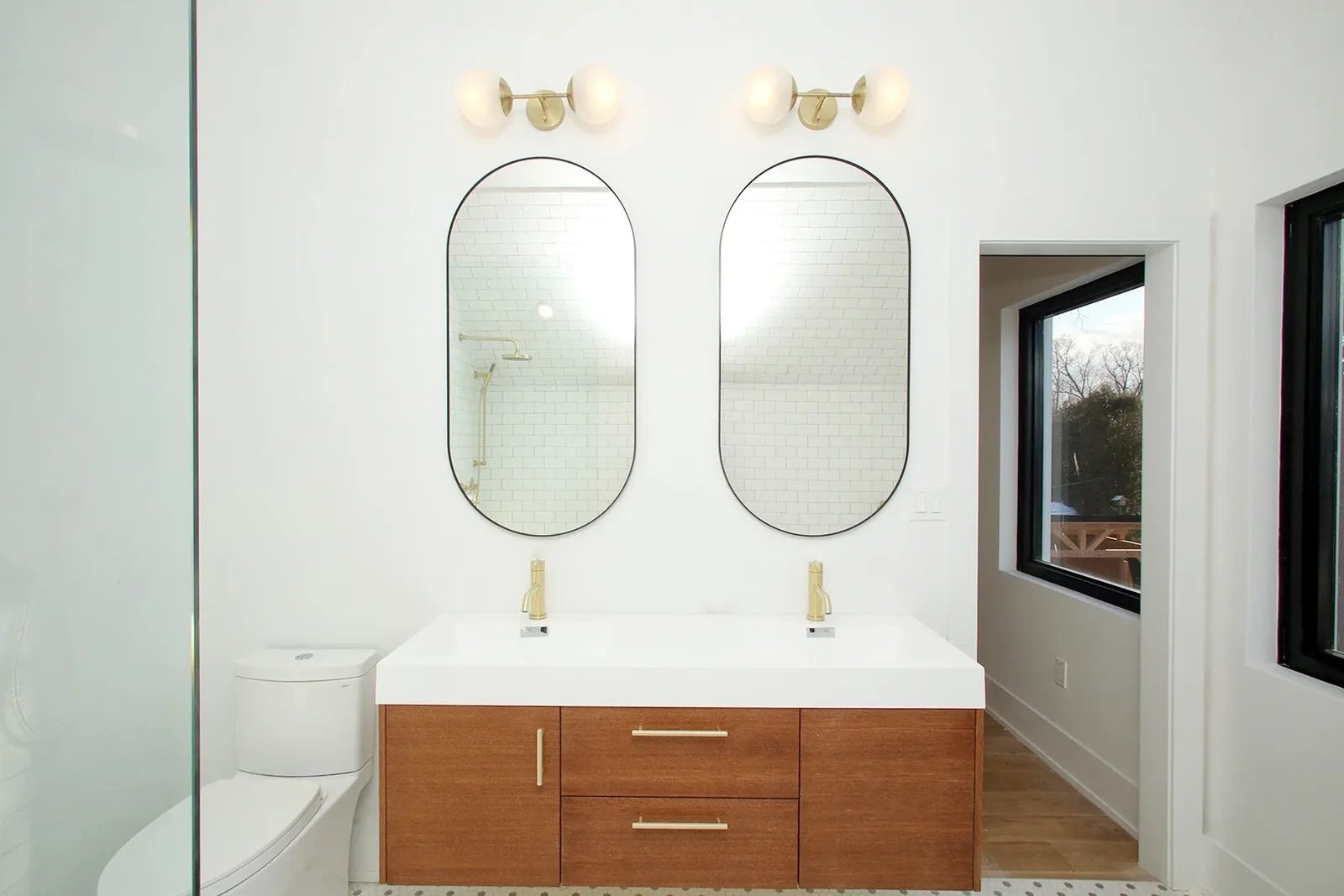 Bright bathroom renovation with a wood double vanity, brass fixtures, arched mirrors, and warm white tile.
