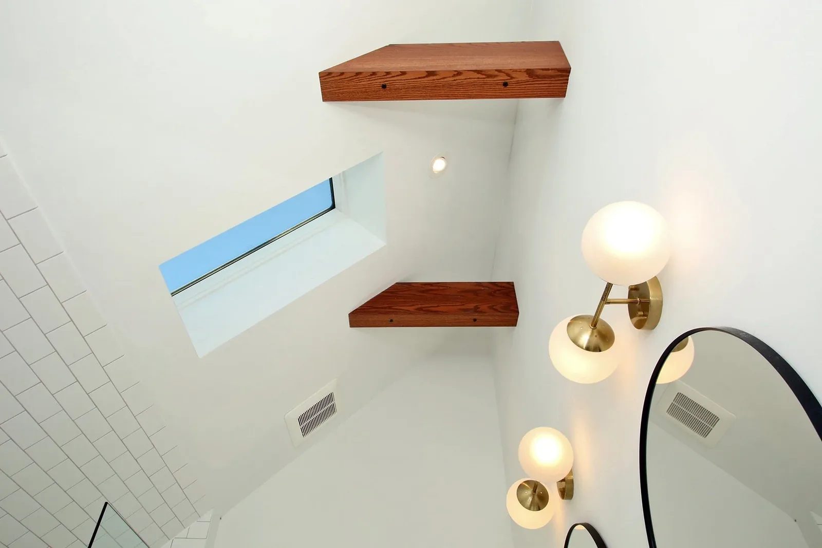 Warm white bathroom detail with a skylight, wood shelves, brass sconces, and a black mirror.