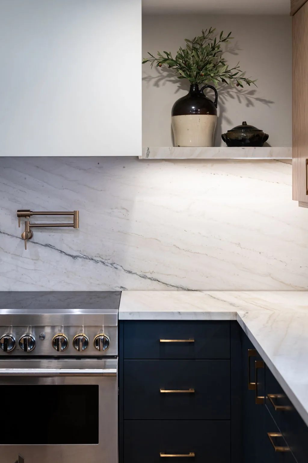 A stainless steel range, brass pot filler and navy drawers sit beneath a white veined stone backsplash and a shelf with ceramics and greenery.