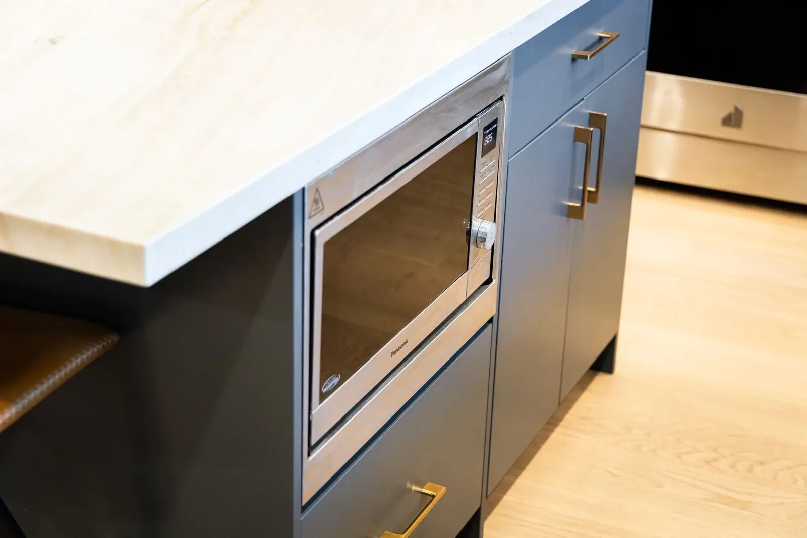 A stainless steel microwave is built into navy island cabinetry with brass pulls beneath a white stone counter.