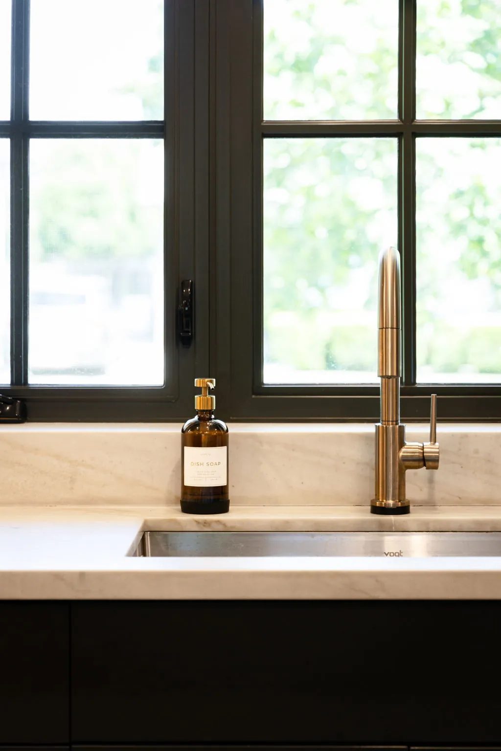 A stainless steel faucet and amber soap dispenser sit beside an undermount sink beneath a black-framed window.