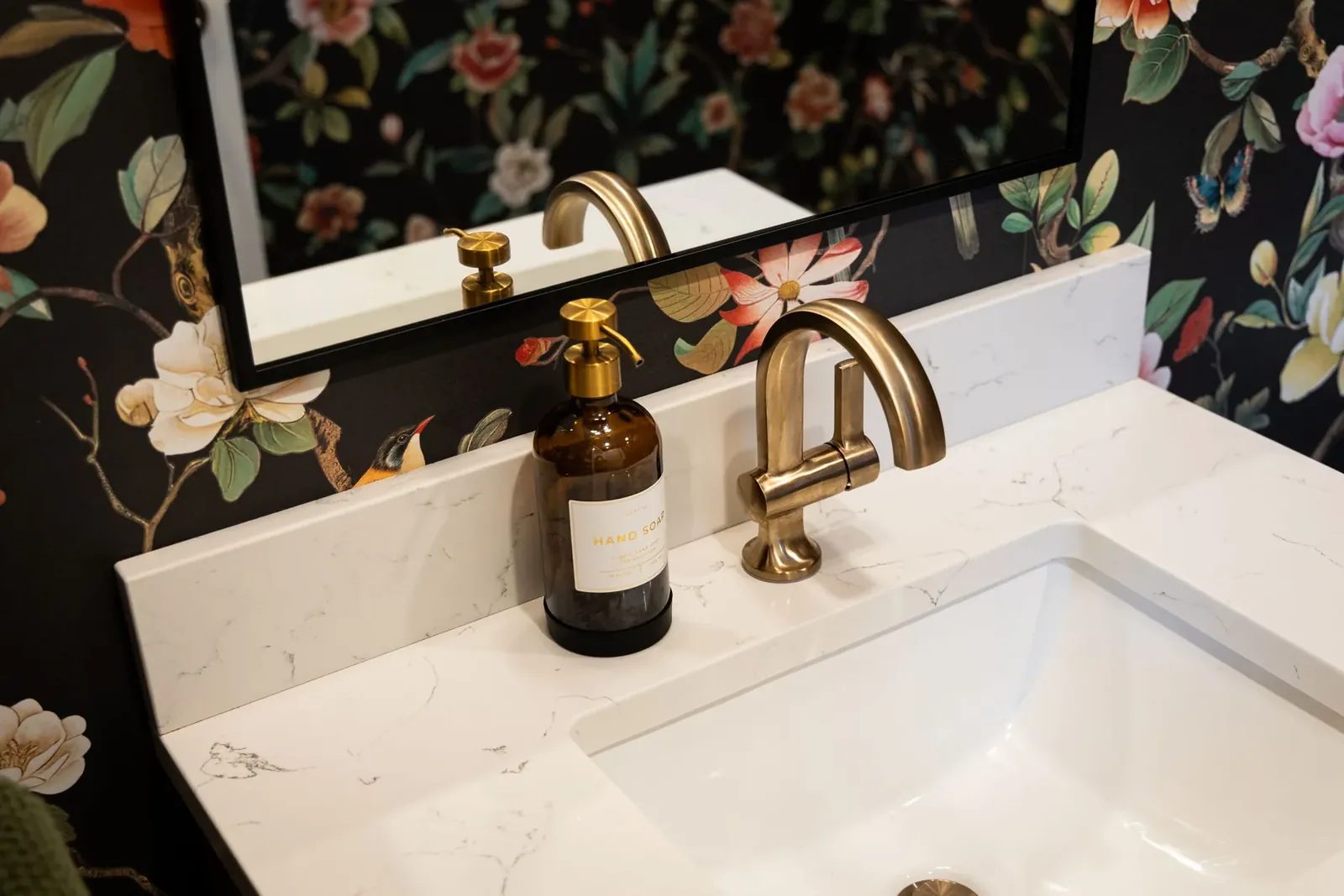 A brushed brass faucet and amber soap dispenser sit on a white veined vanity against black floral wallpaper.
