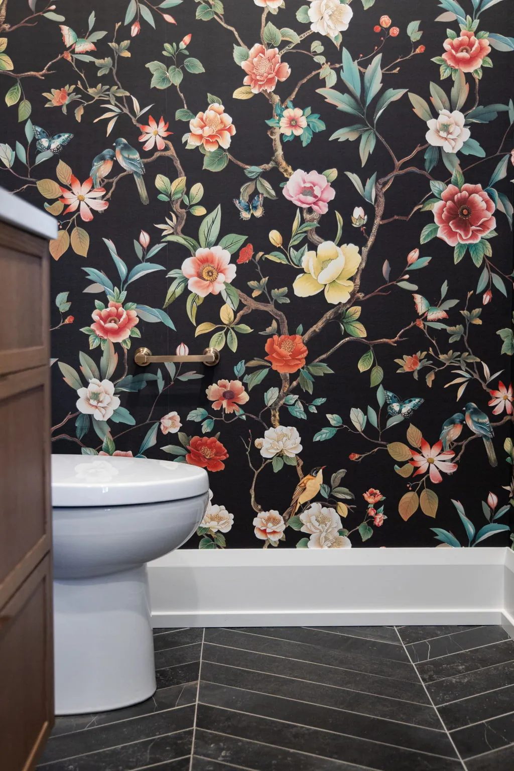 A white toilet and brass towel bar stand against black wallpaper patterned with colourful flowers, birds and butterflies.