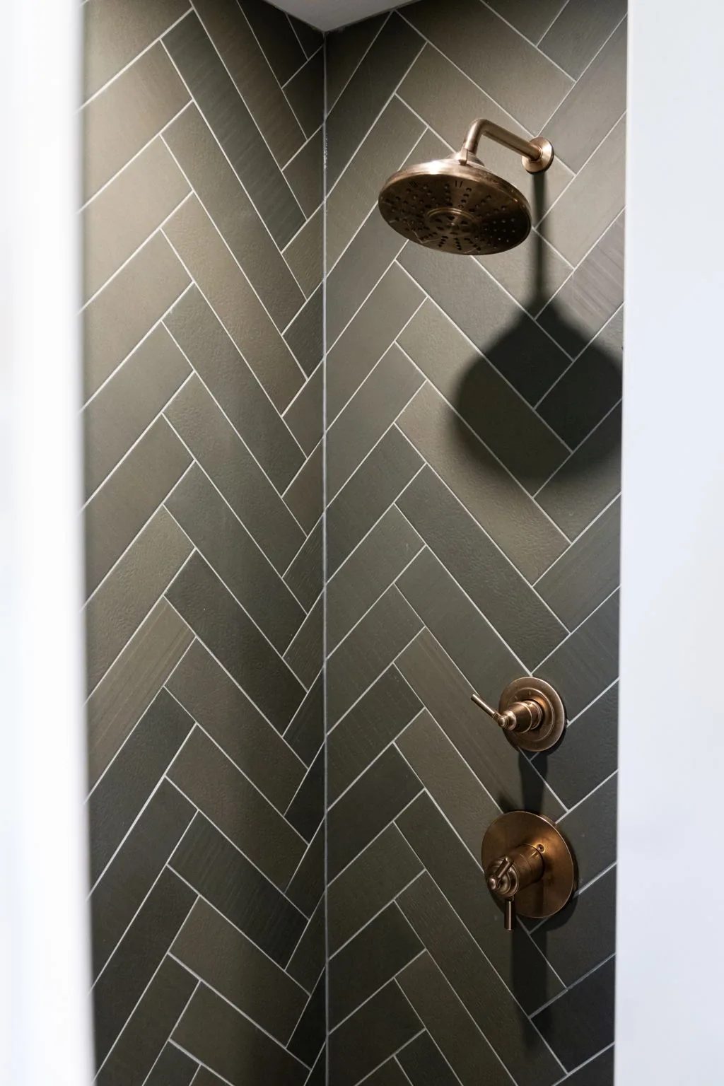 Brass rainfall shower head mounted on a wall of green herringbone tile.