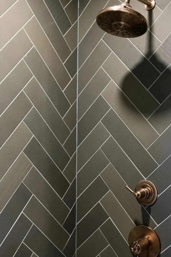 Brushed brass shower fixtures are mounted on dark grey herringbone tile with contrasting light grout.