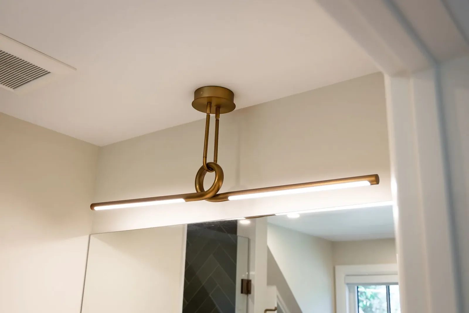 A brass linear light with a central loop hangs above a frameless bathroom mirror.