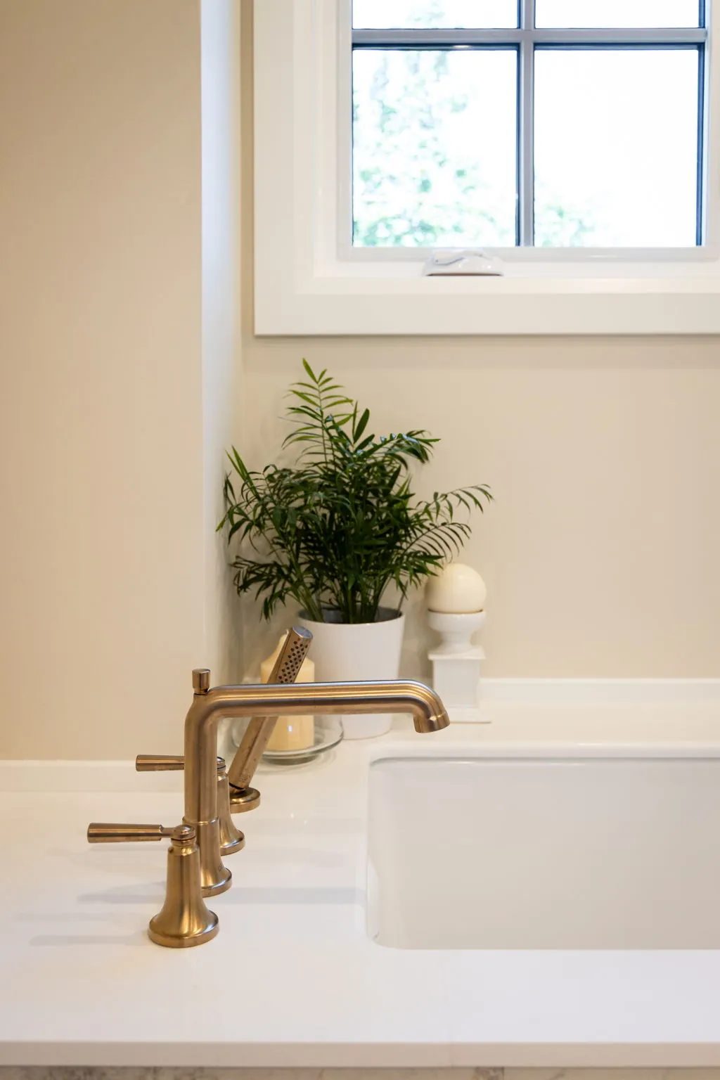 Brushed brass tub fixtures sit on a white surround beside a potted fern and a black-framed window.