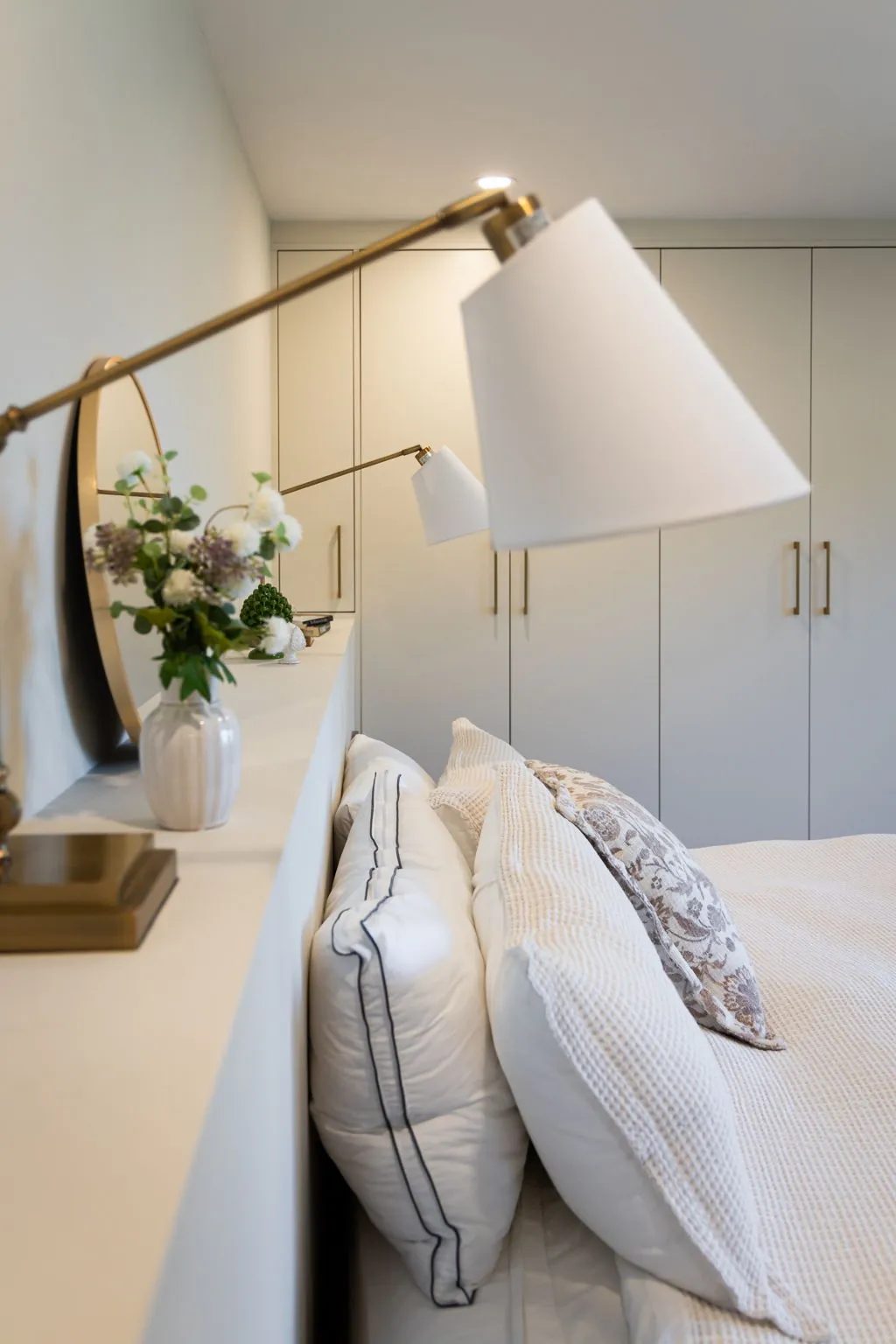 White and patterned pillows rest against a built-in headboard ledge beneath brass reading lamps and white wardrobes.
