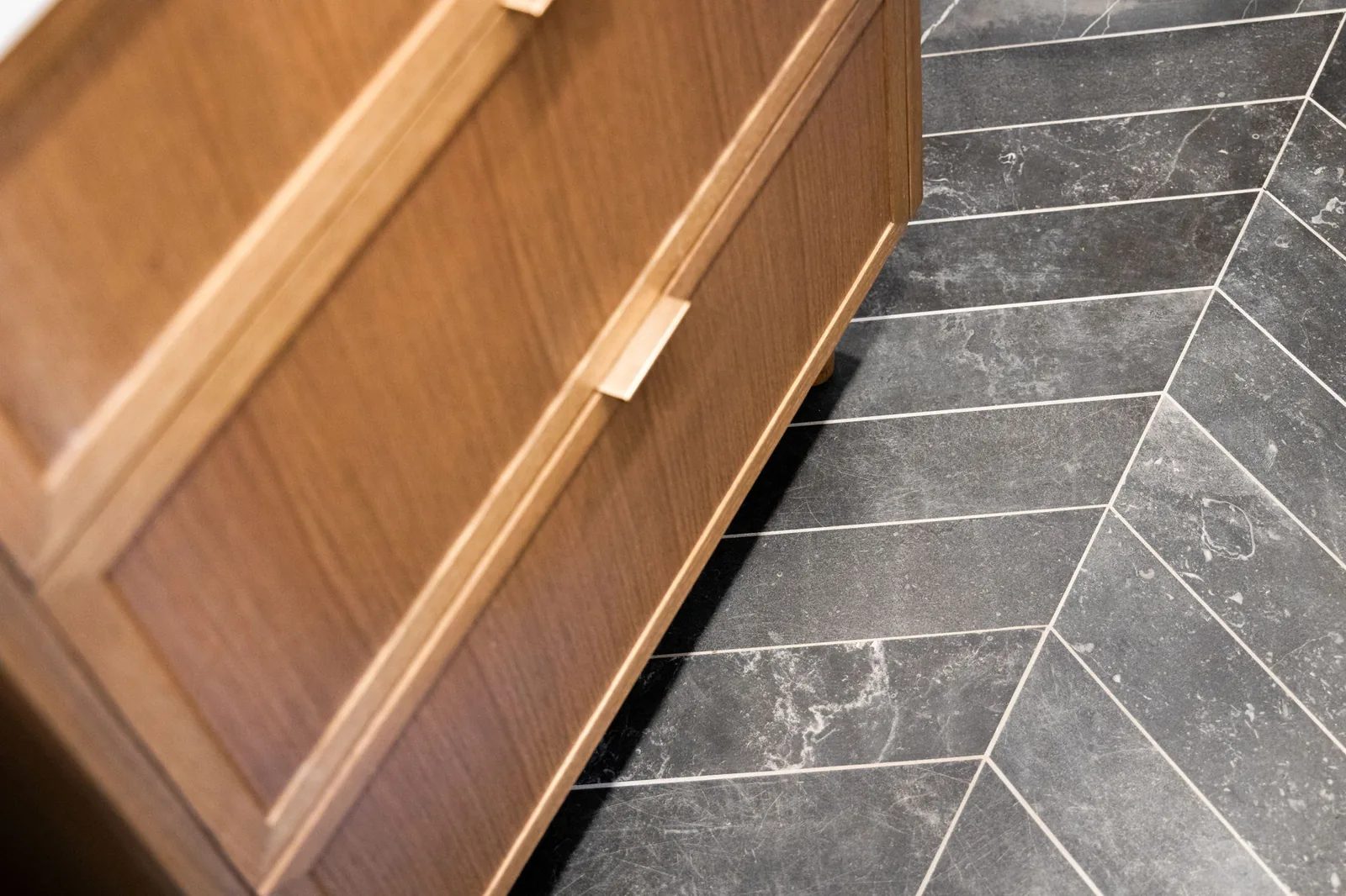 Detail of a walnut baseboard meeting a grey herringbone stone tile floor.