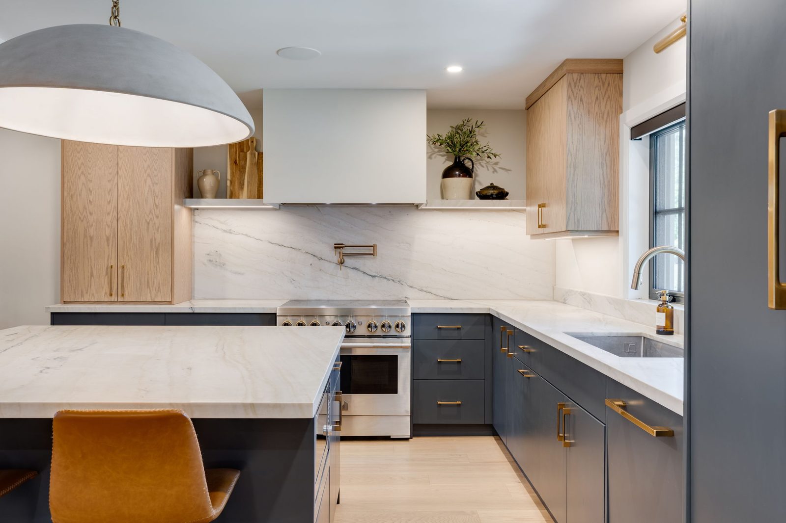 Wide view across the quartzite waterfall island to the navy base cabinetry, the white oak uppers and the stone slab backsplash beyond.