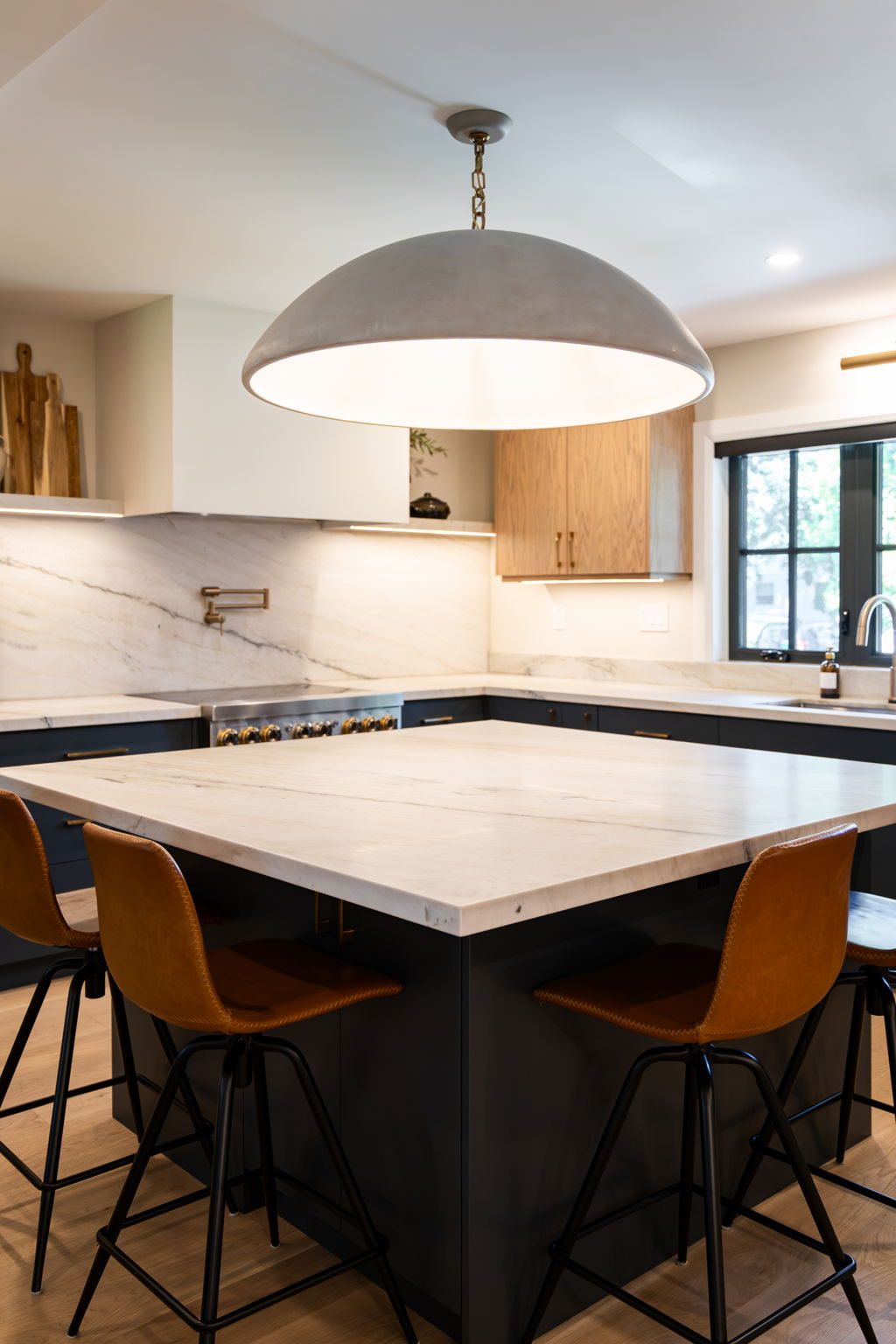 Quartzite waterfall island under a wide dome pendant, with amber leather stools tucked in and white oak and navy cabinetry filling the walls behind.