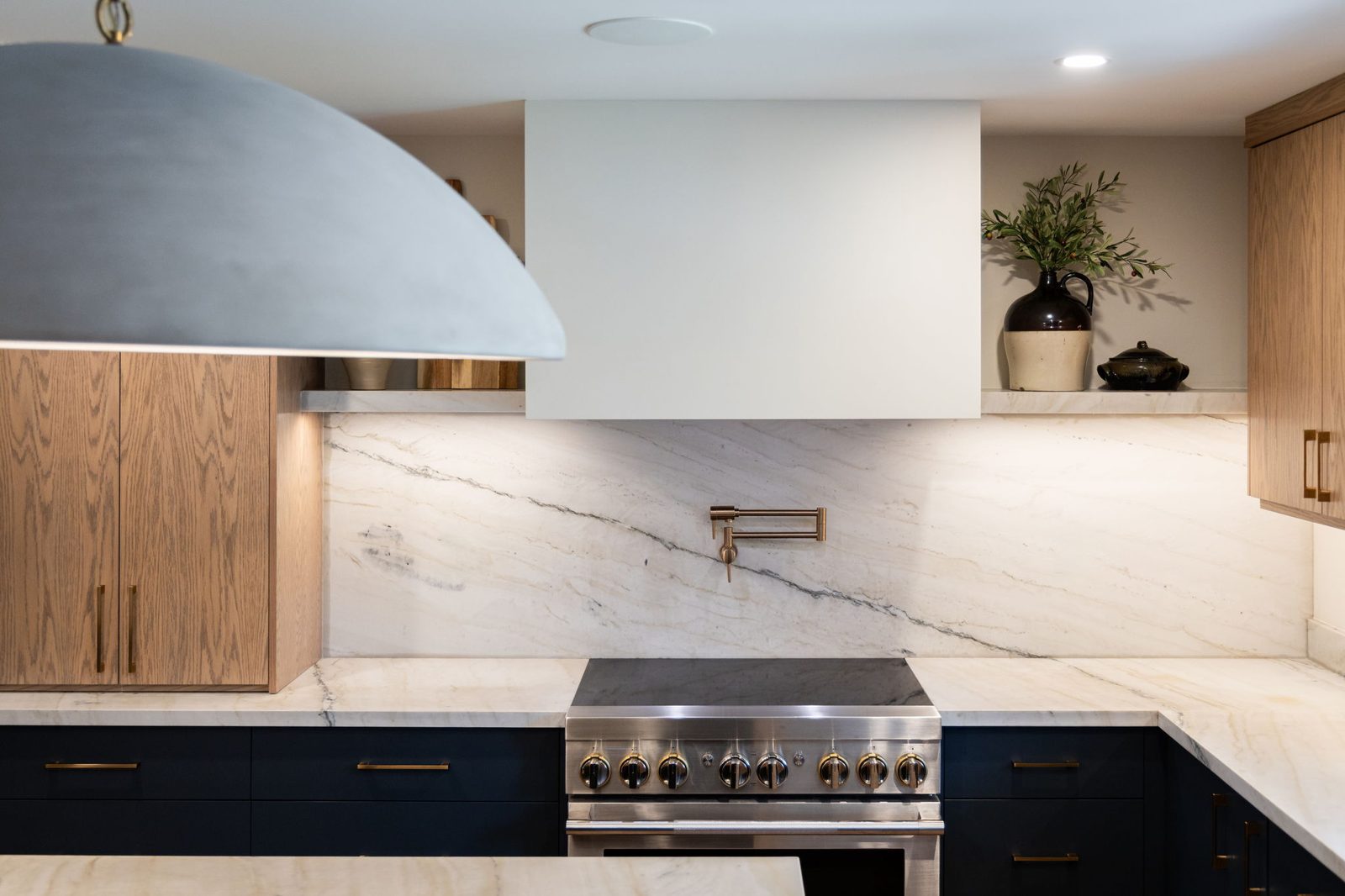 Range wall in full, with a bookmatched stone slab backsplash, a plaster hood, white oak cabinetry, a brass pot filler and a stainless pro range.