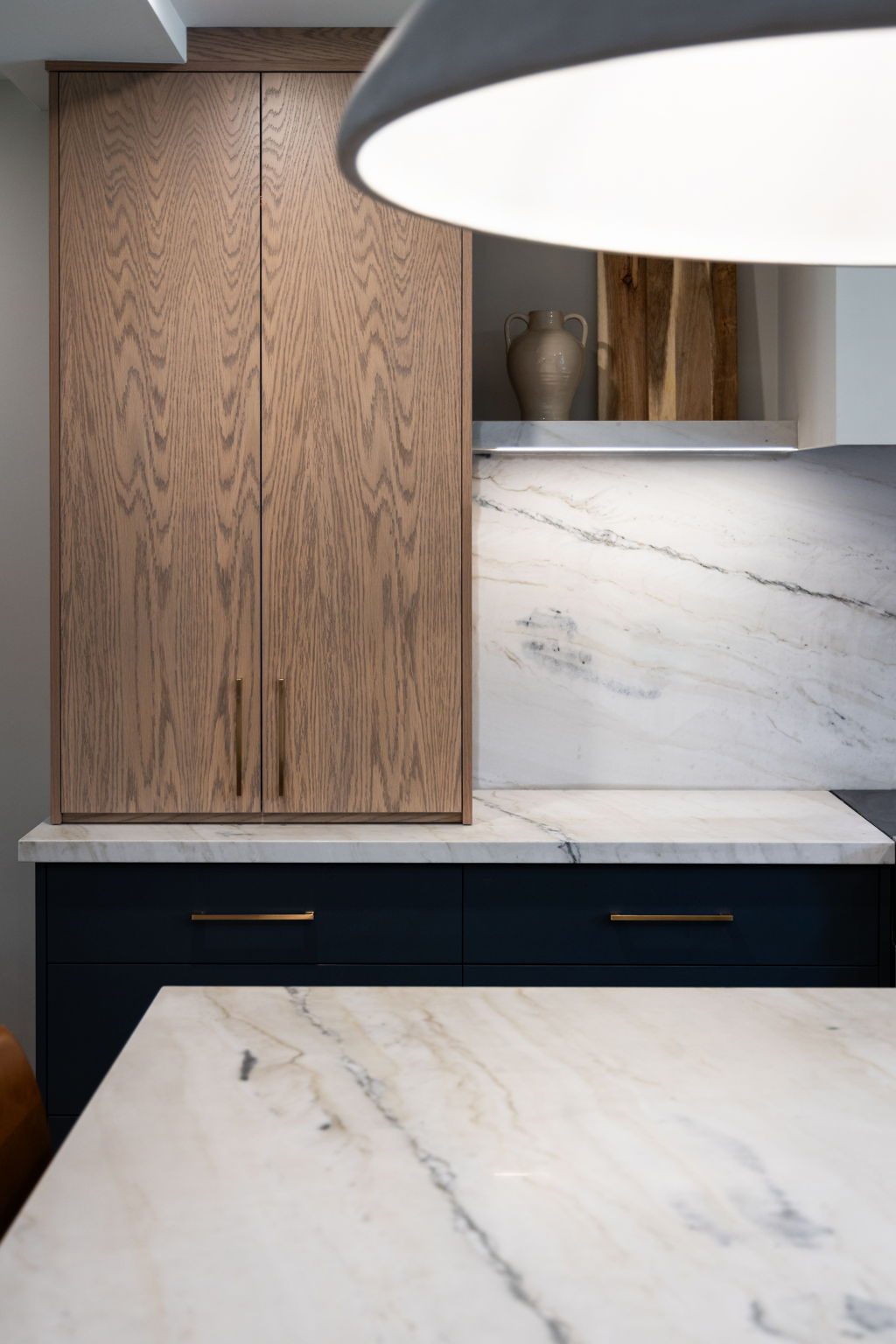 Full-height white oak cabinet set against the veined stone slab, with navy base cabinets and brass bar pulls below.