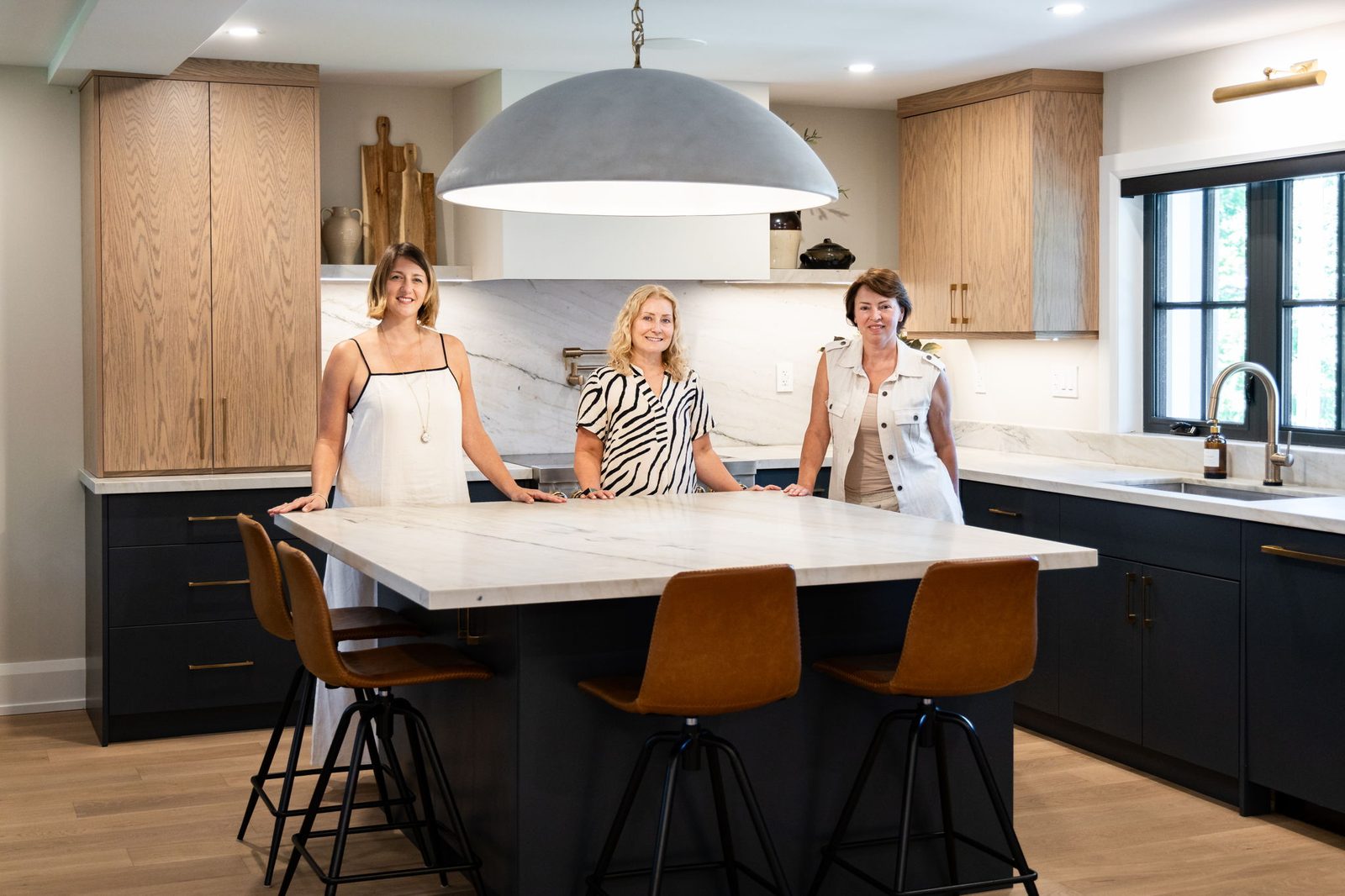 Three homeowners standing together at the quartzite island beneath the dome pendant, with the white oak and navy kitchen cabinetry behind them.