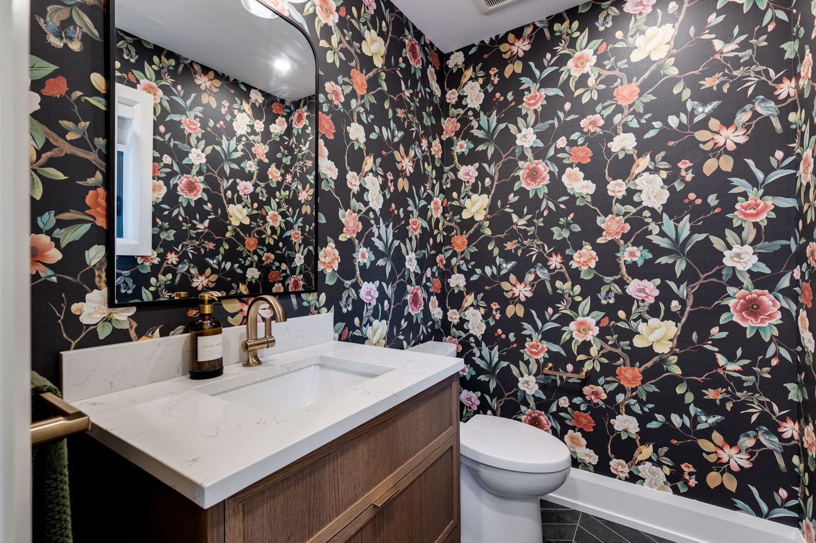 Powder room with dark floral wallpaper, a floating walnut vanity, a stone counter with a mitred return and a brass gooseneck faucet.