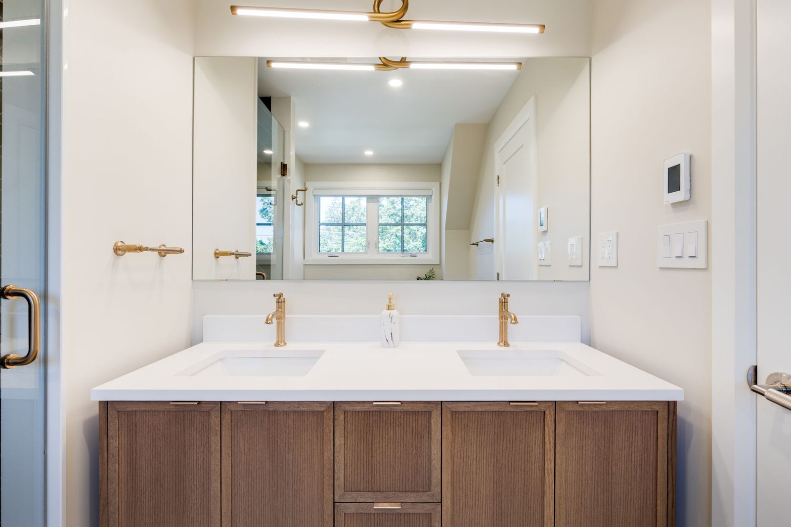 Reeded walnut double vanity straight on, with twin brass faucets, a stone counter and a full-width mirror under a linear brass light.