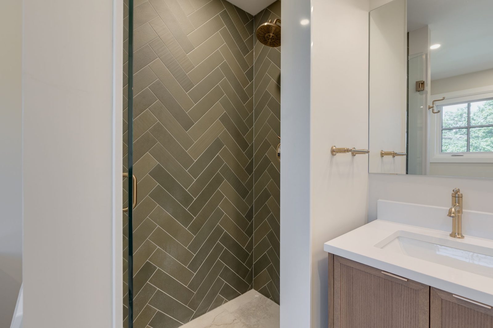 Curbless walk-in shower clad in dark green herringbone tile behind a frameless glass panel, with the walnut vanity beyond.