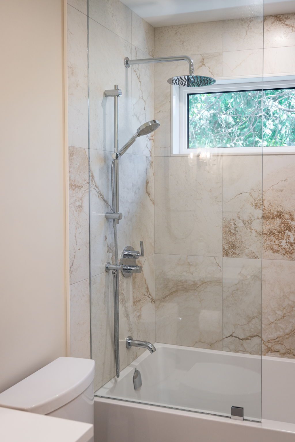 Tub and shower alcove lined in large-format stone-look tile, with a chrome riser, a rainfall head and a glass screen.