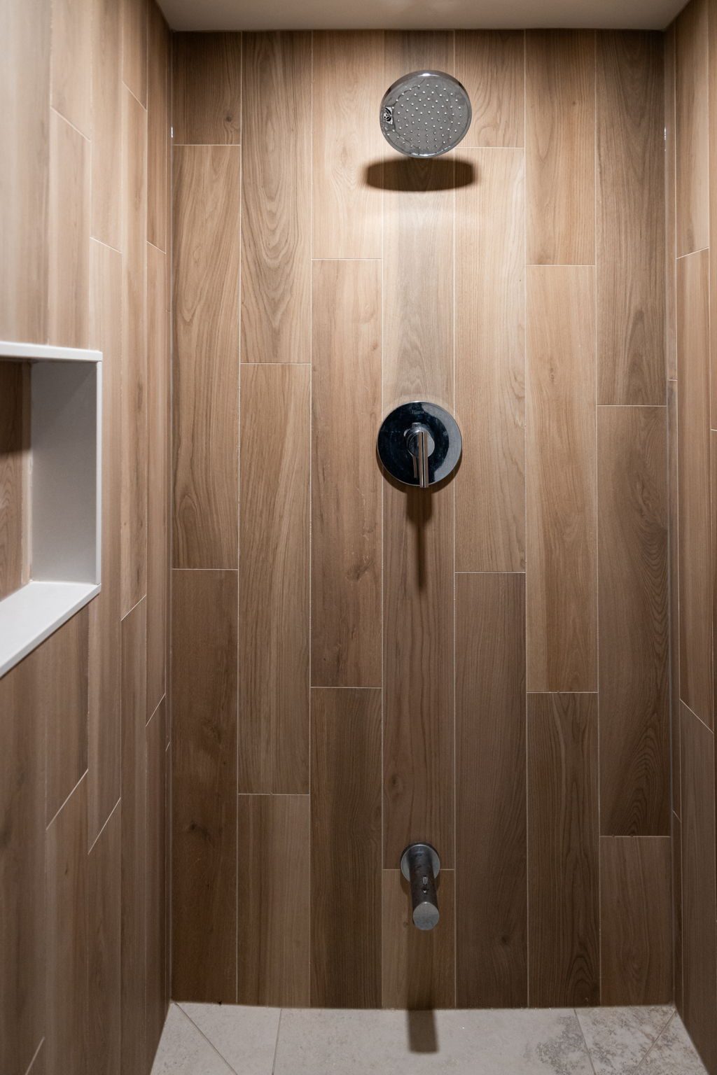 Wood-look tile shower straight on, with a chrome rainfall head, a wall valve and a recessed niche.